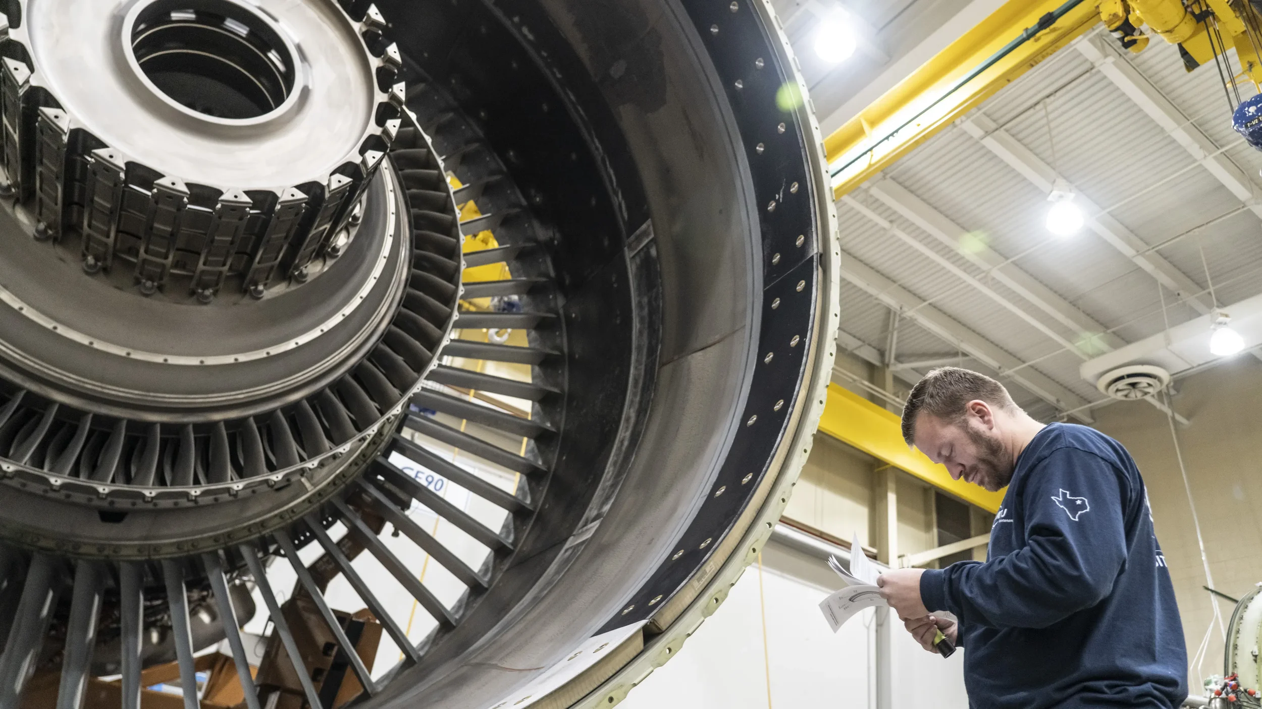 Dallas industrial photography capturing a technician reviewing documentation while inspecting a large jet engine inside an aviation maintenance facility, highlighting quality control, compliance, and technical accuracy.