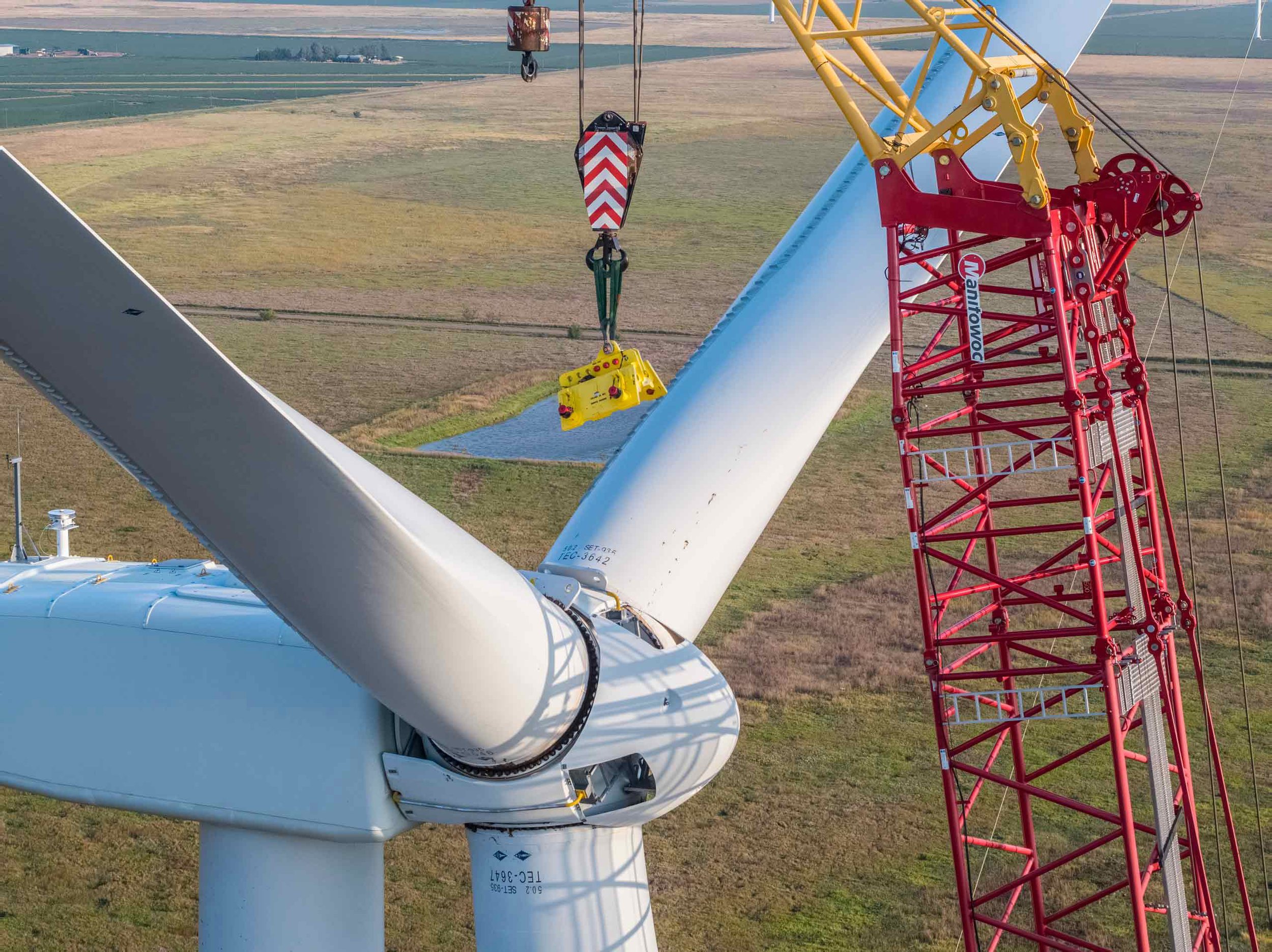Aerial drone photography capturing wind turbine blade installation using a heavy-lift crane, highlighting precision engineering, renewable energy infrastructure, and large-scale industrial construction in an active wind farm environment.