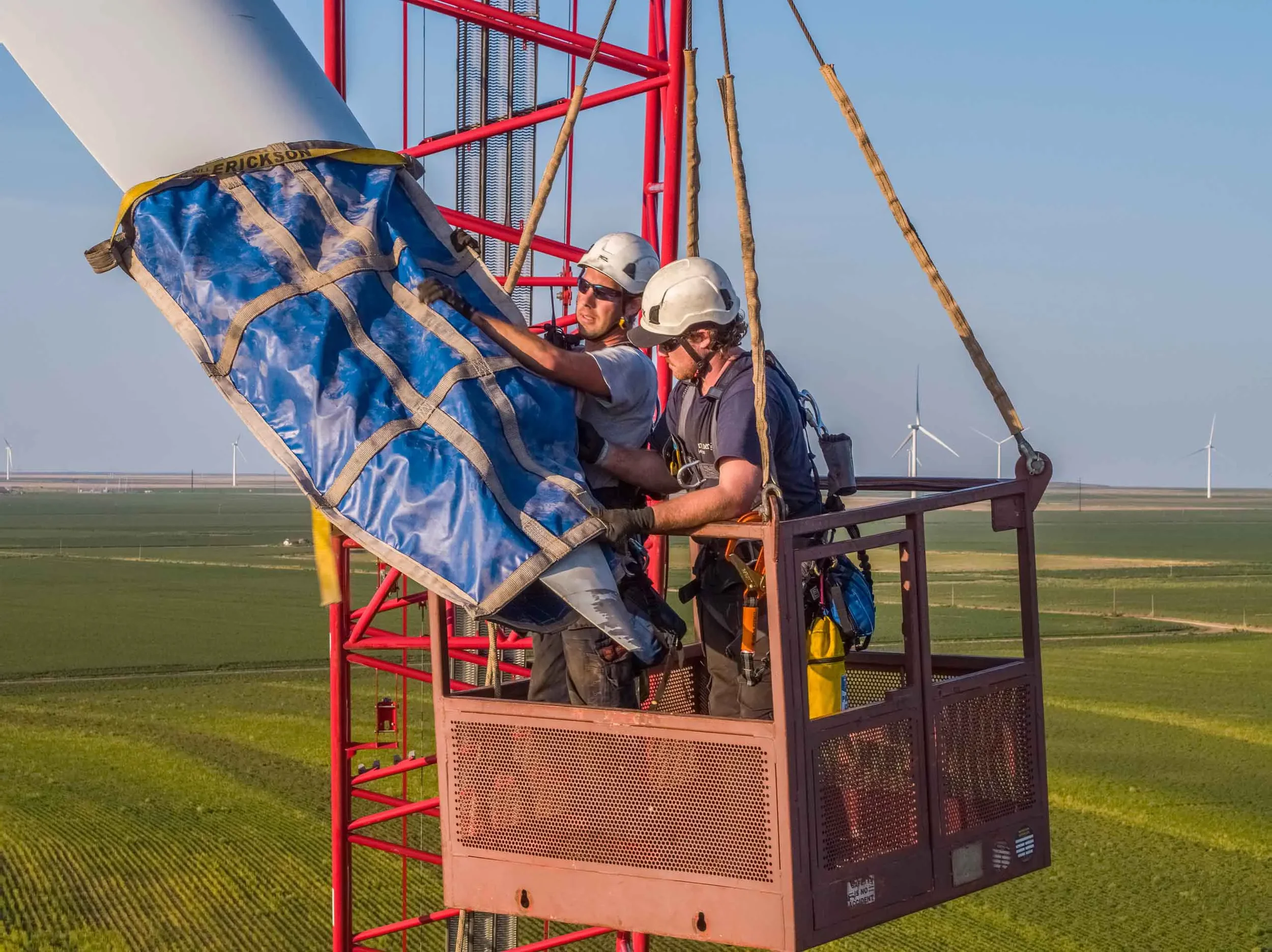 Professional drone photography documenting wind turbine blade installation using a crane basket, emphasizing industrial safety, renewable energy construction, and complex lift operations captured from above.