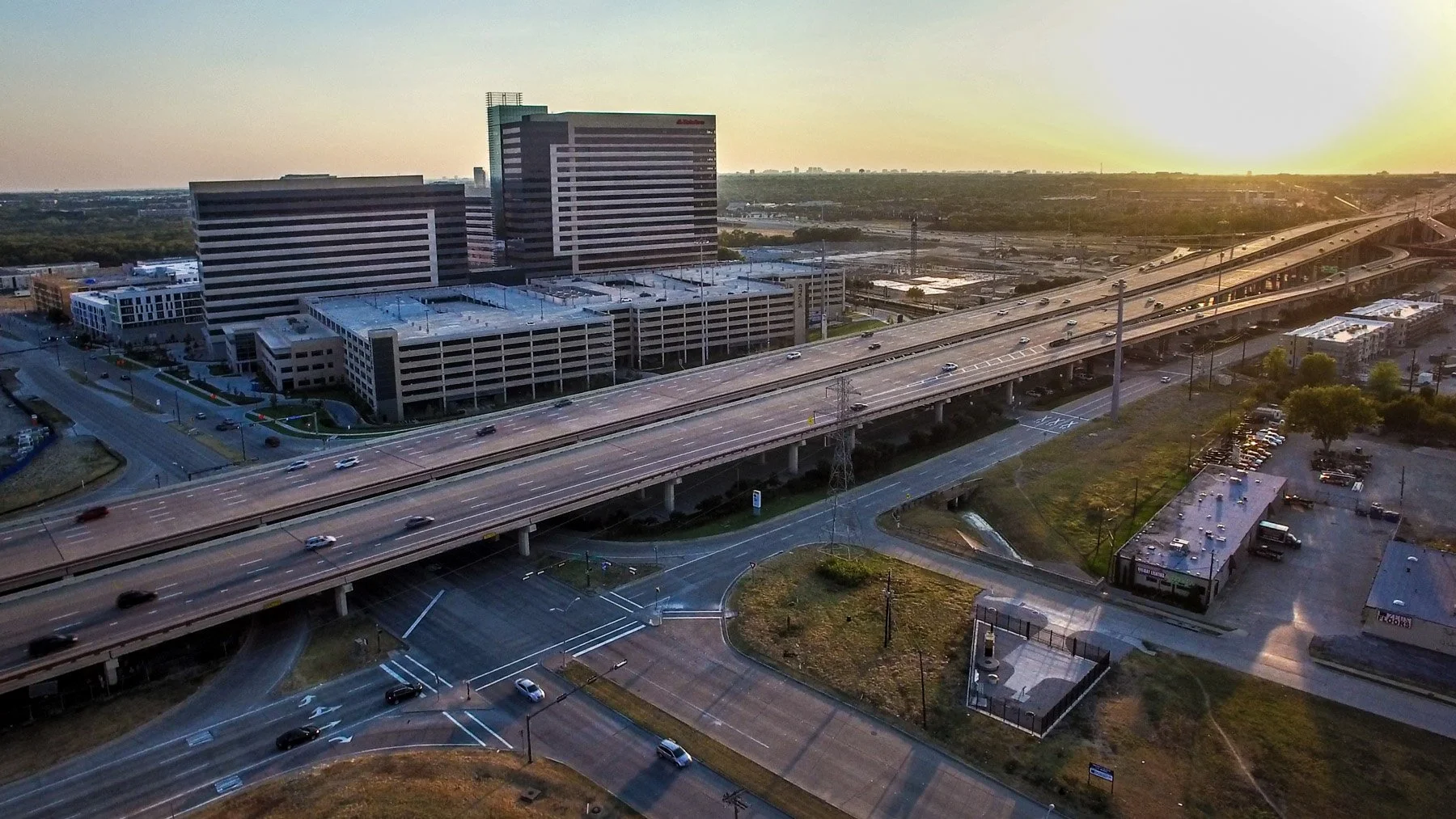 Drone photography capturing a major Dallas highway interchange at golden hour, highlighting transportation infrastructure, traffic flow, and surrounding commercial development.
