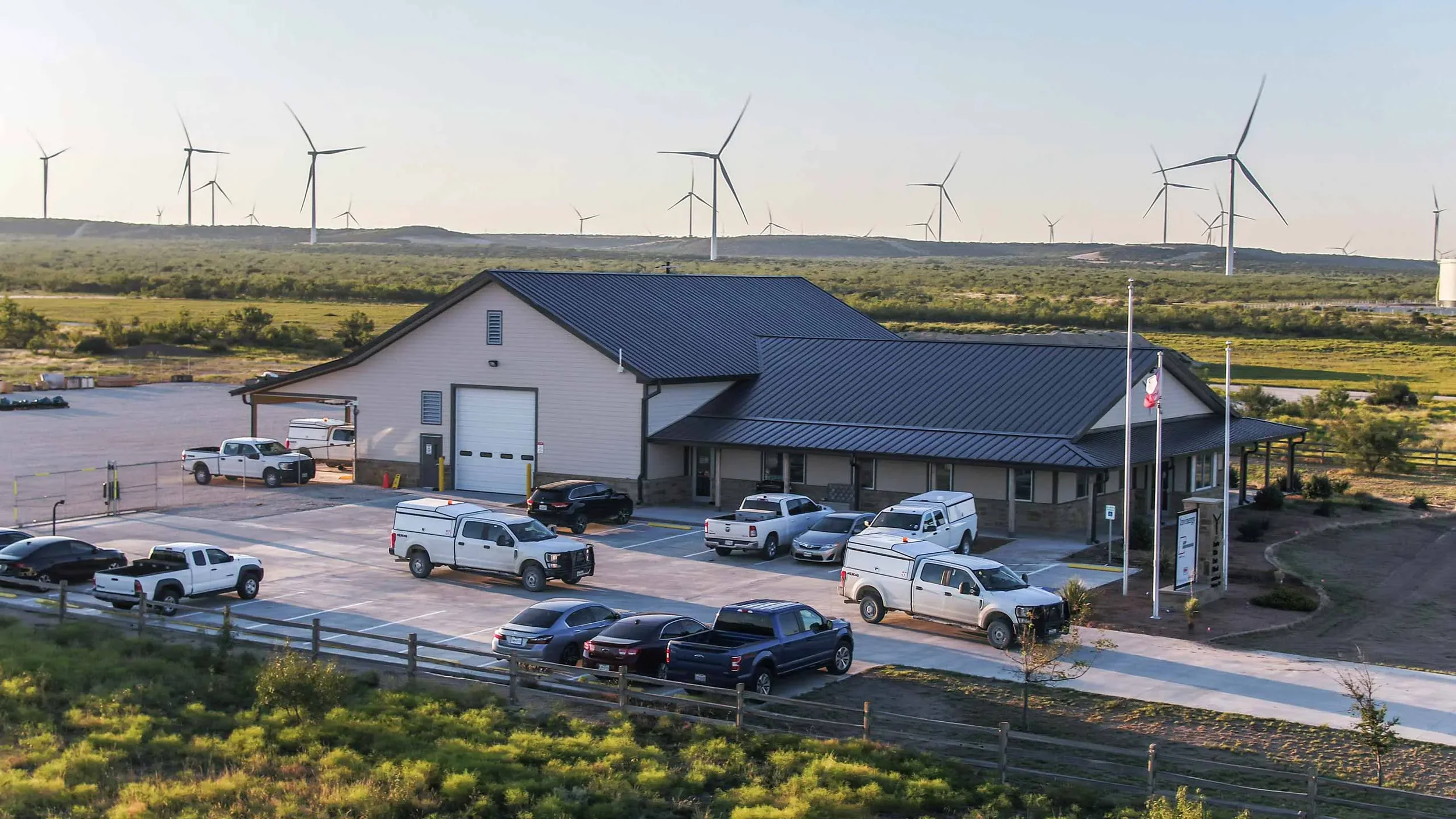 Drone aerial image of a wind energy operations facility surrounded by active wind turbines, showcasing renewable energy infrastructure, utility support buildings, and industrial operations photography in Texas.