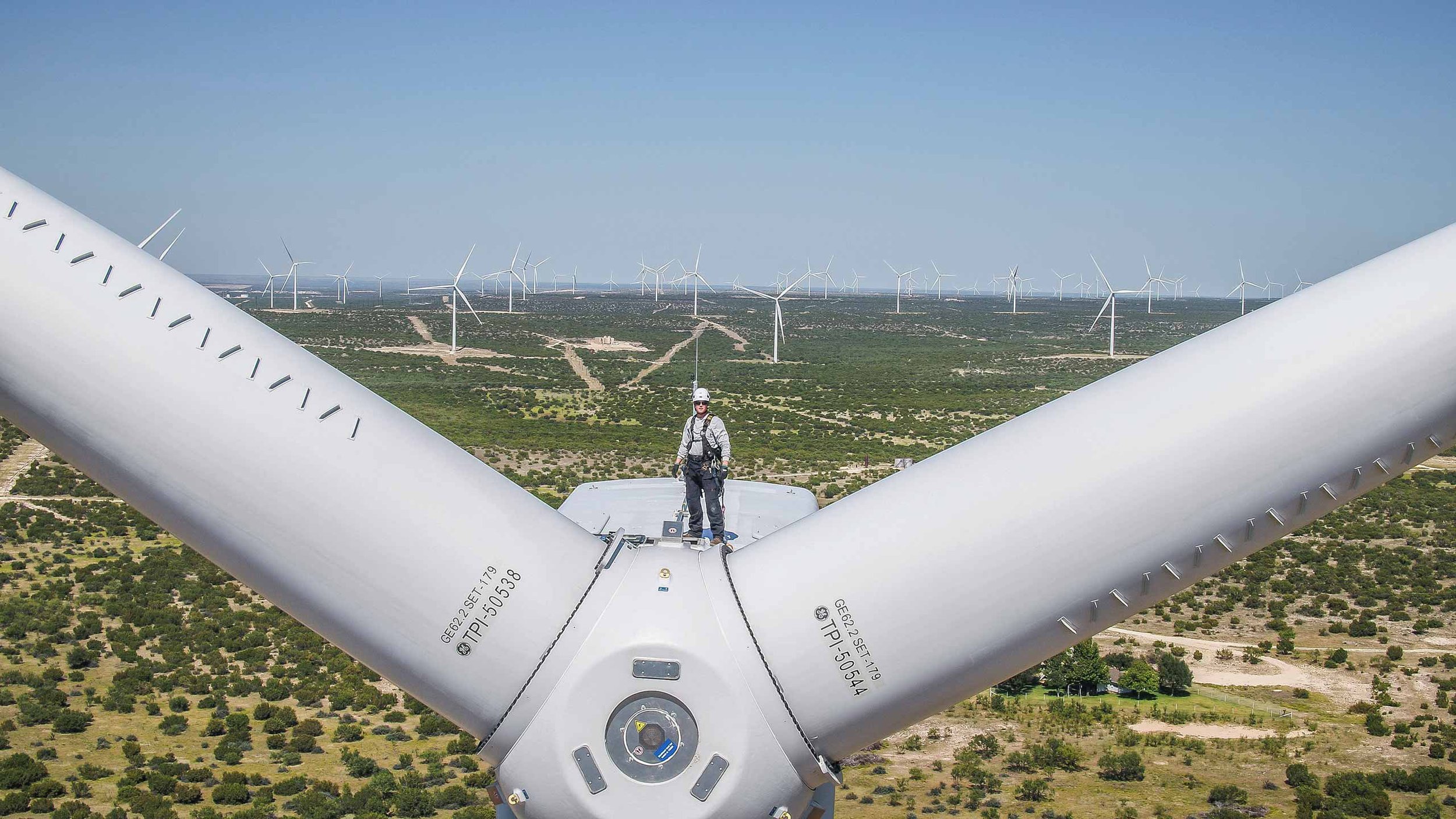 High-altitude drone photograph of a wind turbine technician standing atop a turbine hub, with a vast wind farm in the background, showcasing renewable energy infrastructure and safety-focused industrial operations.