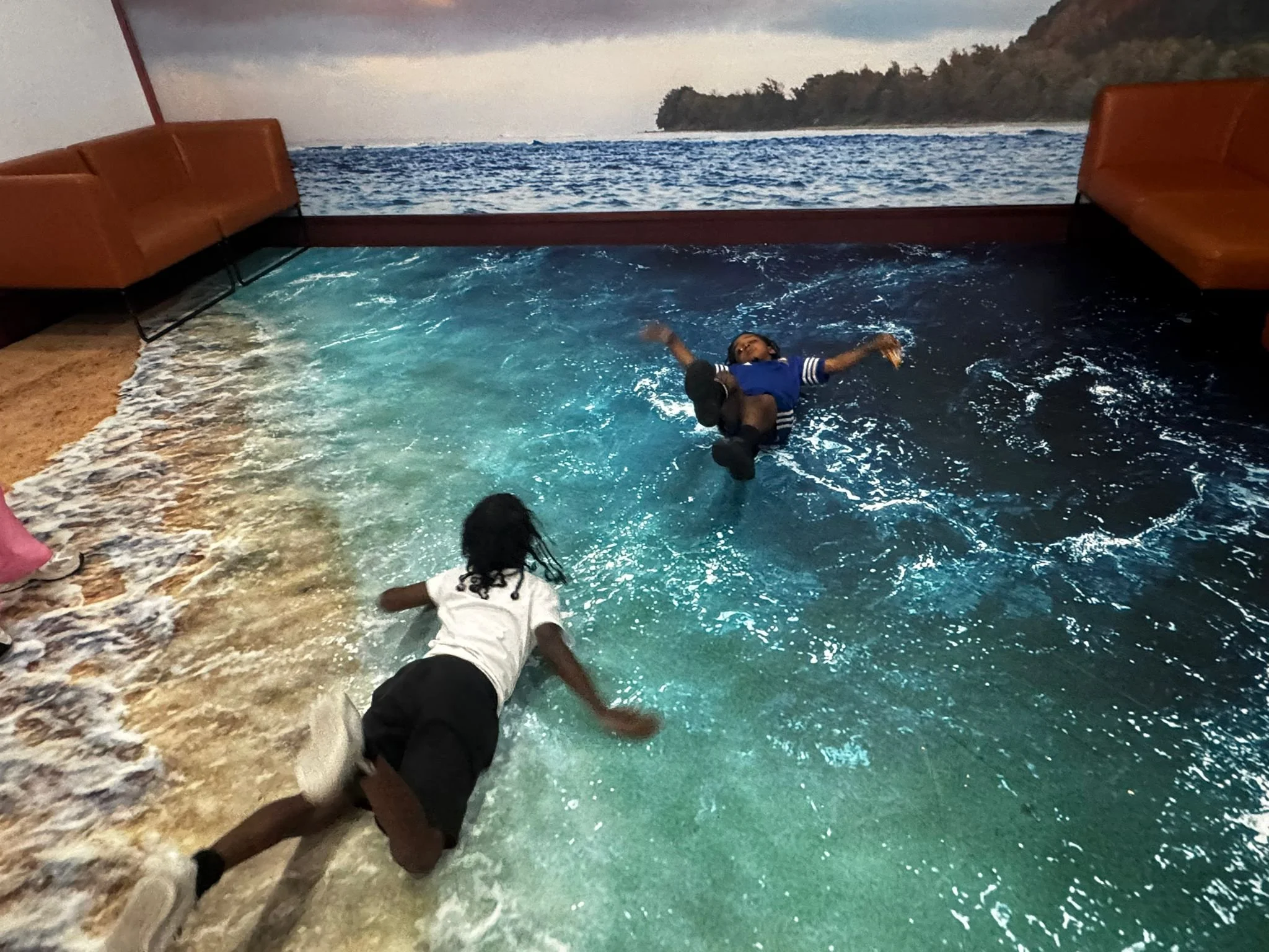 Two children are playing in a room with a floor that looks like a beach and water, with a backdrop of ocean scenery. One child is crawling near the foreground, and the other is lying on the floor, both with the water-themed design.