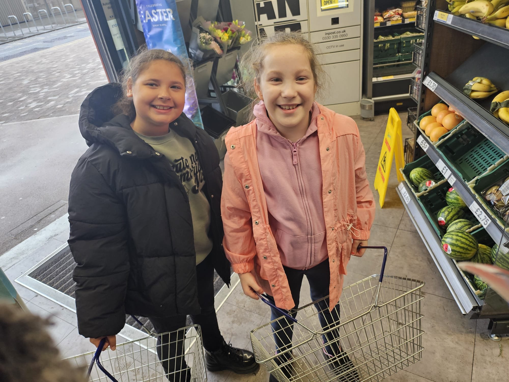 Two young girls with shopping baskets inside a grocery store, smiling at the camera, with produce like watermelons and bananas on shelves behind them.