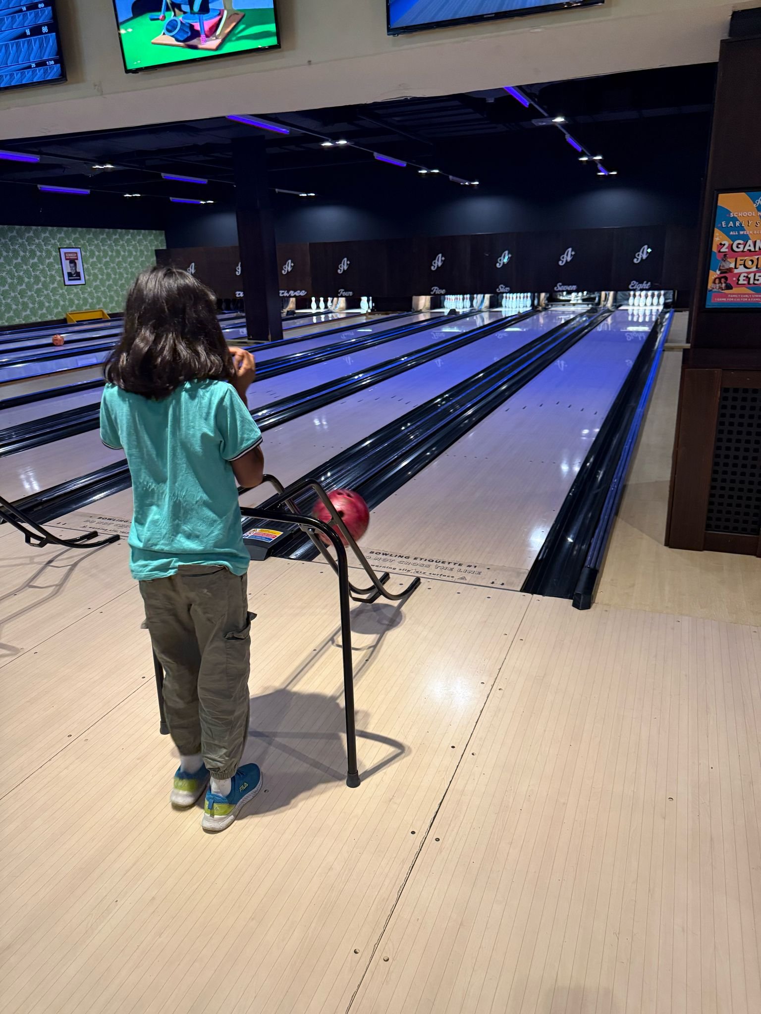 A young girl with dark hair, wearing a turquoise shirt and beige pants, stands on a bowling alley lane about to roll a bowling ball.