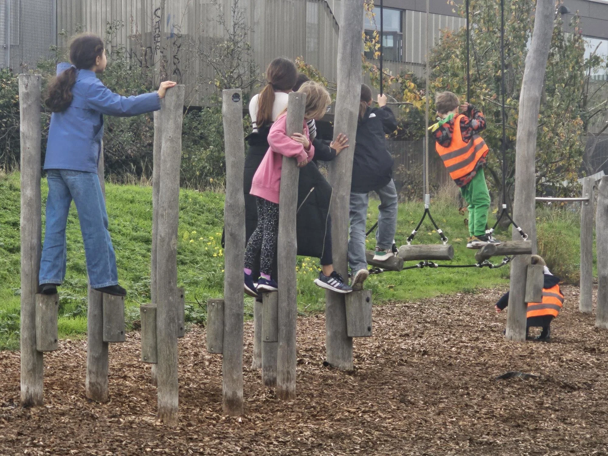 Children playing on wooden playground equipment with logs and suspension bridges in an outdoor park area, some wearing colorful jackets, with trees and buildings in the background.