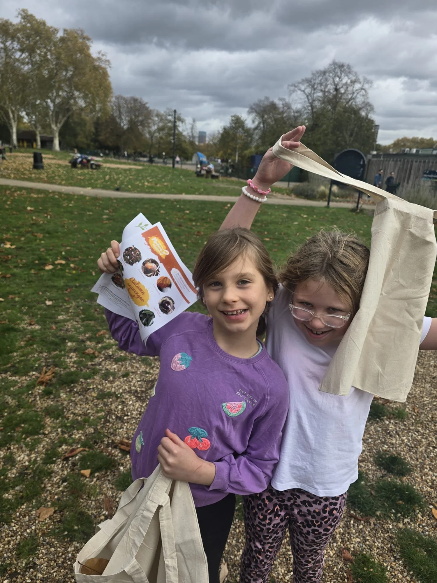 Two girls smiling and holding reusable tote bags outdoors in a park on a cloudy day, one girl holding a flyer with images of muffins.