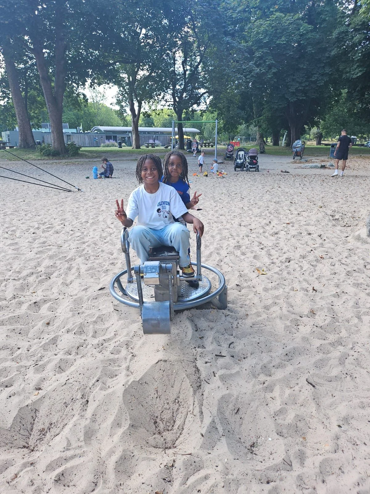 Two children, a girl and a boy, are playing on a merry-go-round at a sandy playground. The girl is sitting in front, smiling and making peace signs with both hands. The background shows other children playing, trees, and a person walking.