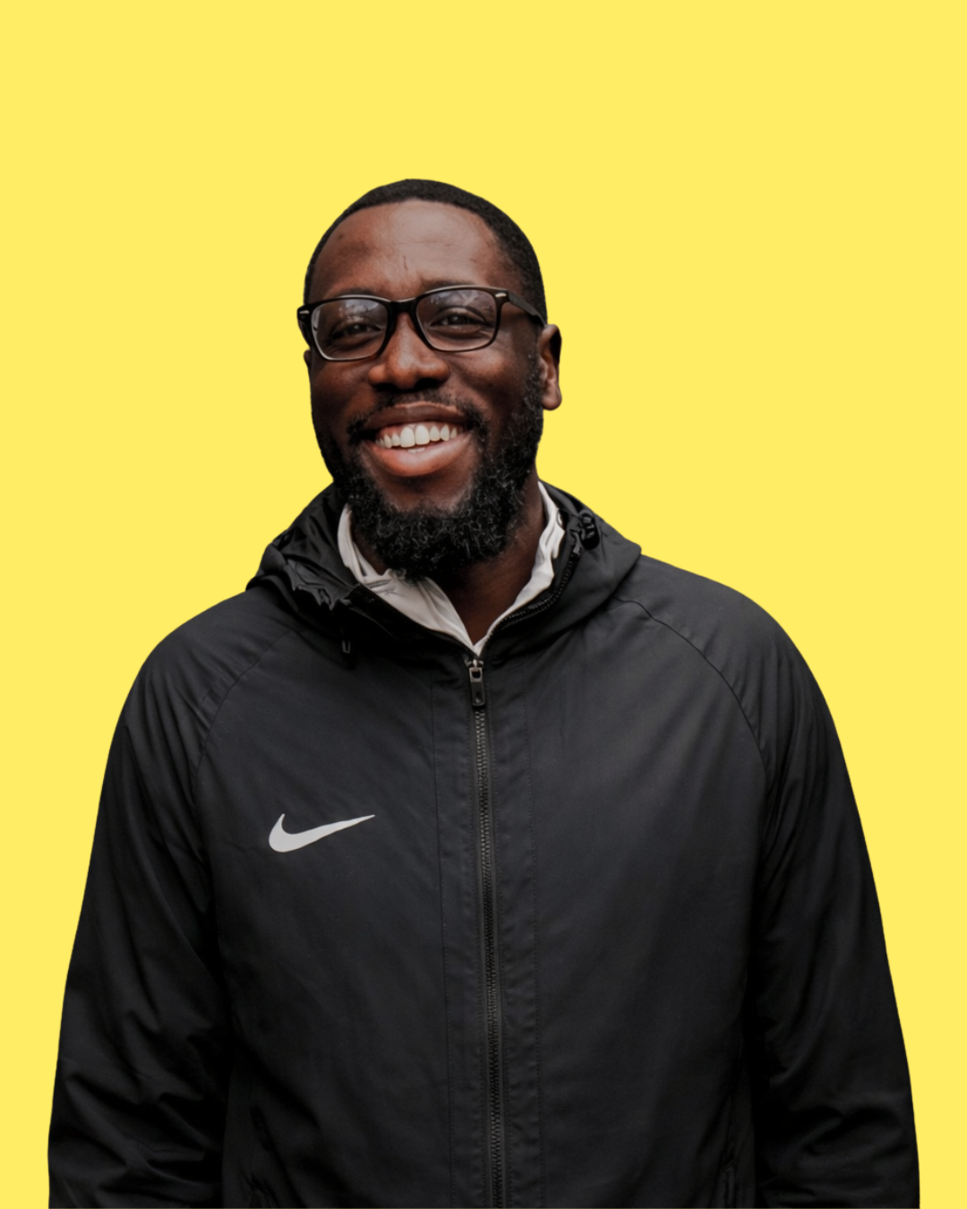 A smiling man wearing glasses and a black Nike jacket against a yellow background.