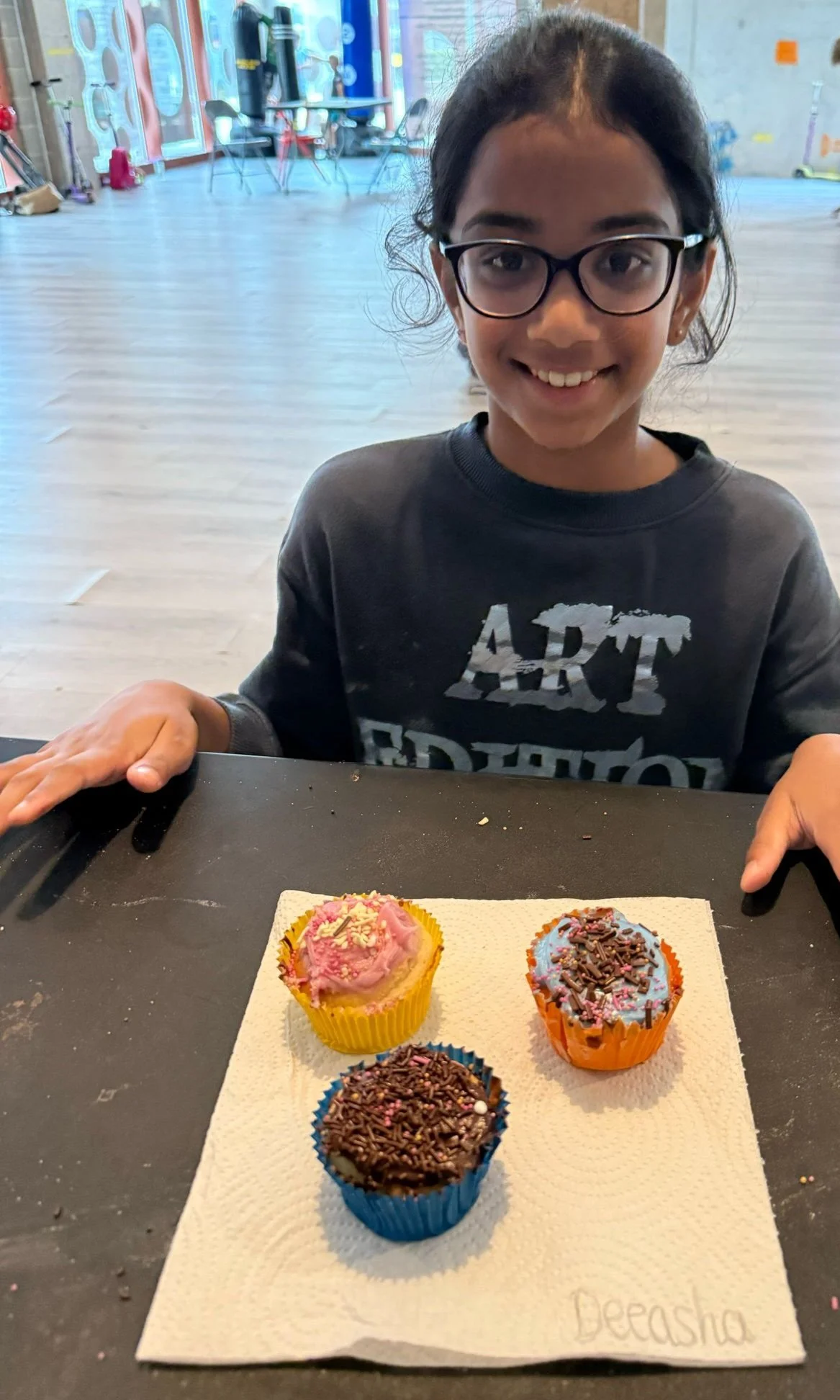 A young girl with glasses smiling at a table with three cupcakes decorated with pink and chocolate frosting and sprinkles, placed on a paper towel in a classroom or activity space.