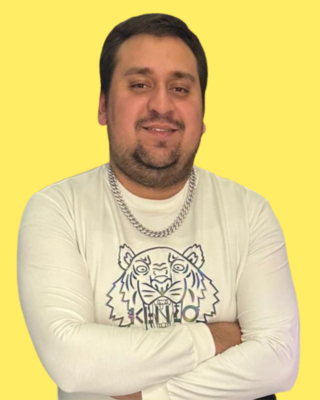 A man with short dark hair, a beard, and a mustache, smiling, wearing a white long sleeve shirt with a lion graphic, crossed arms, yellow background.