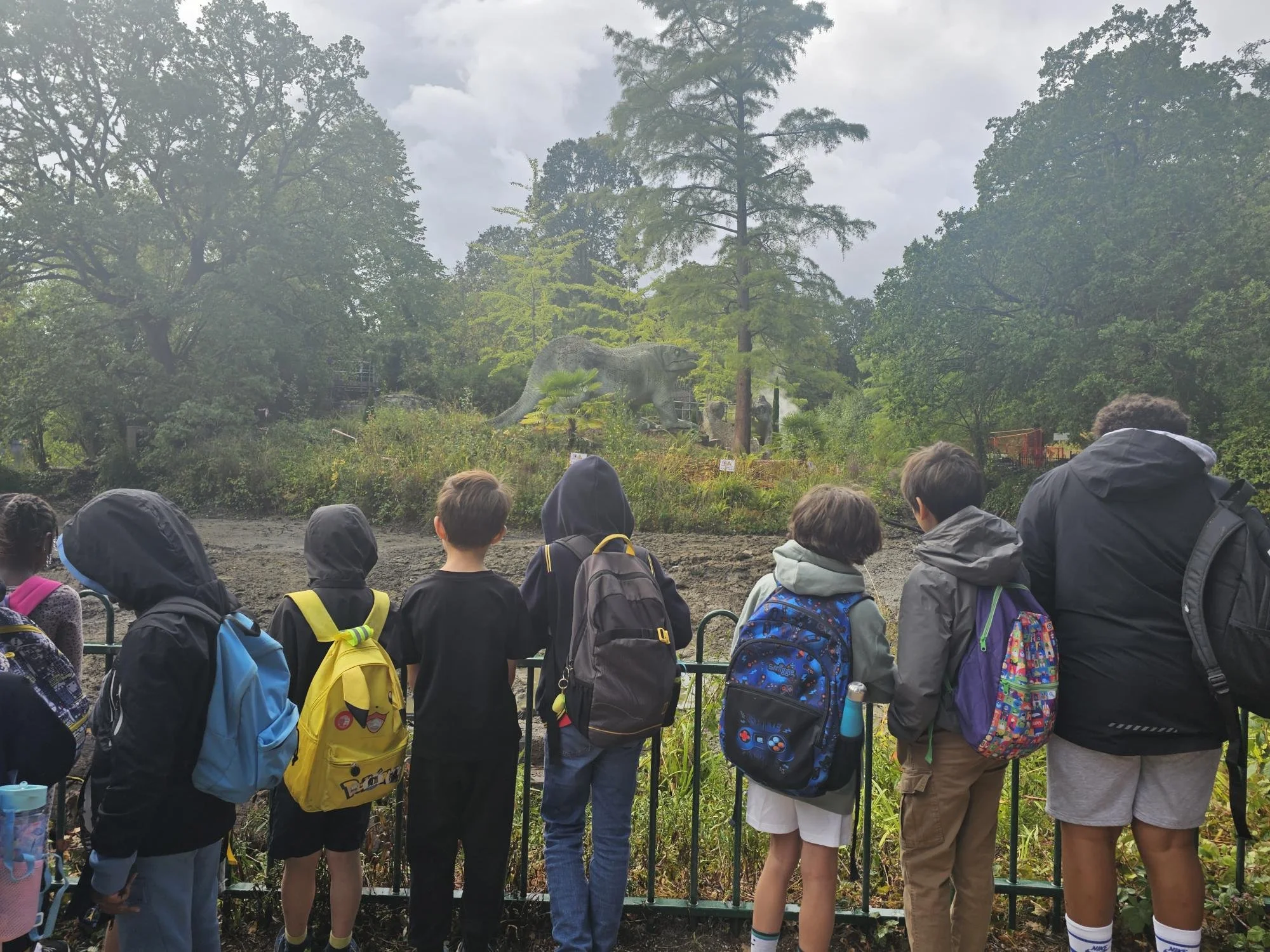 Group of children and adults observing a dinosaur exhibit at a zoo or museum outdoor area, with trees and cloudy sky in the background.