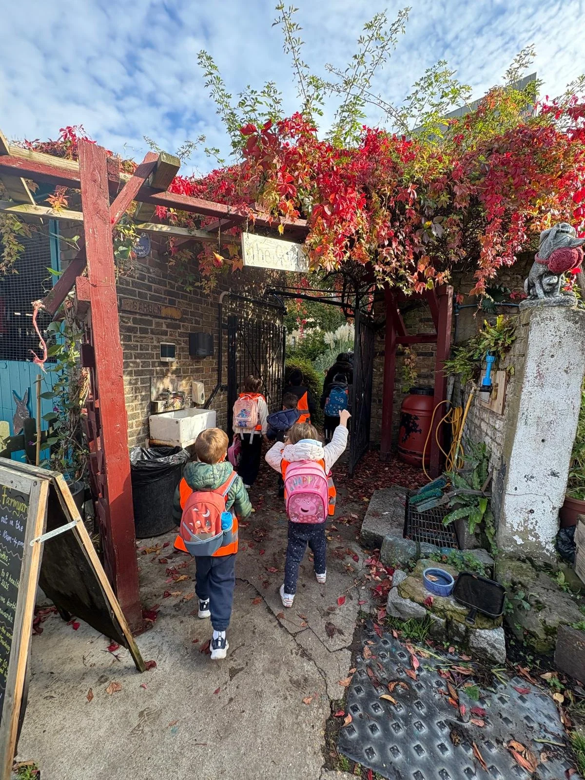 A group of children with backpacks walking through a garden gate with red and green foliage, on a paved path under a partly cloudy sky.