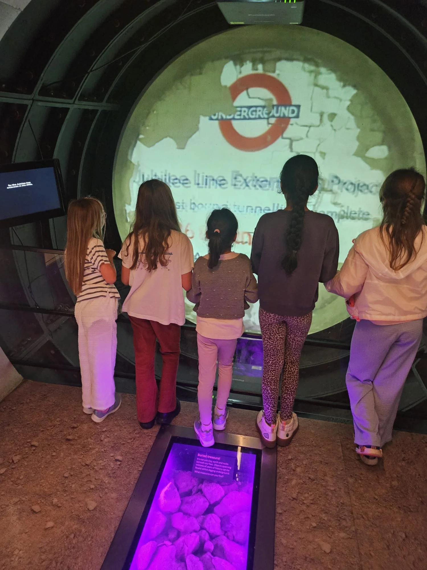 Five children standing in front of an interactive exhibit about the London Underground, which features a large circular display with the Underground logo and text. There is a purple-lit display on the floor showing rocks or fossils.