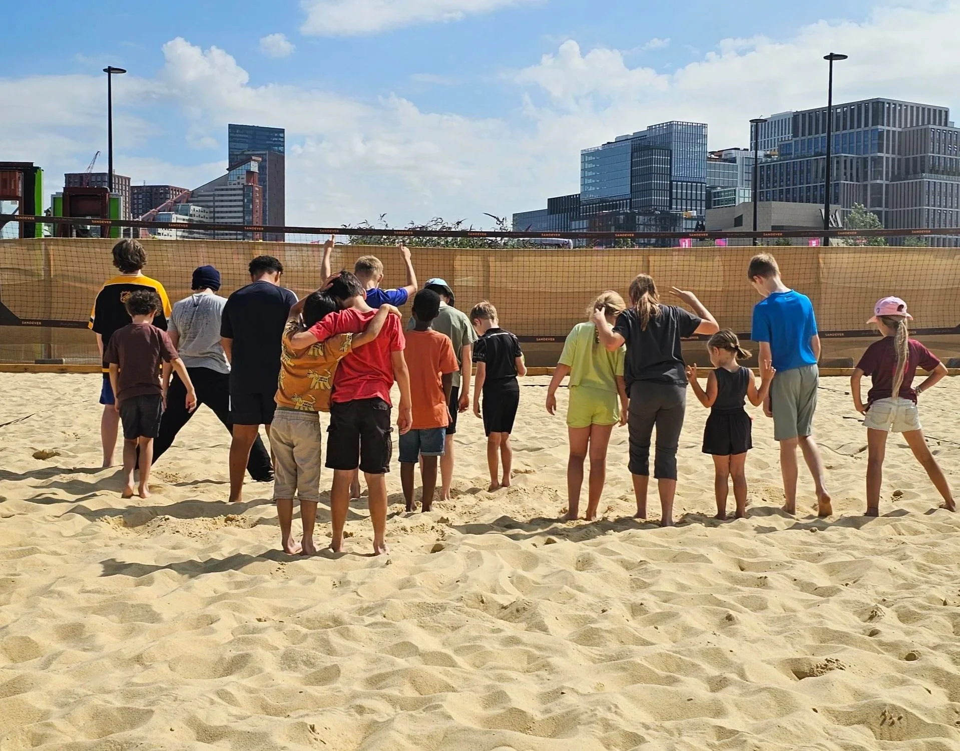 Group of children standing on sandy beach volleyball court, some embracing, others talking, with tall city buildings and blue sky in the background.