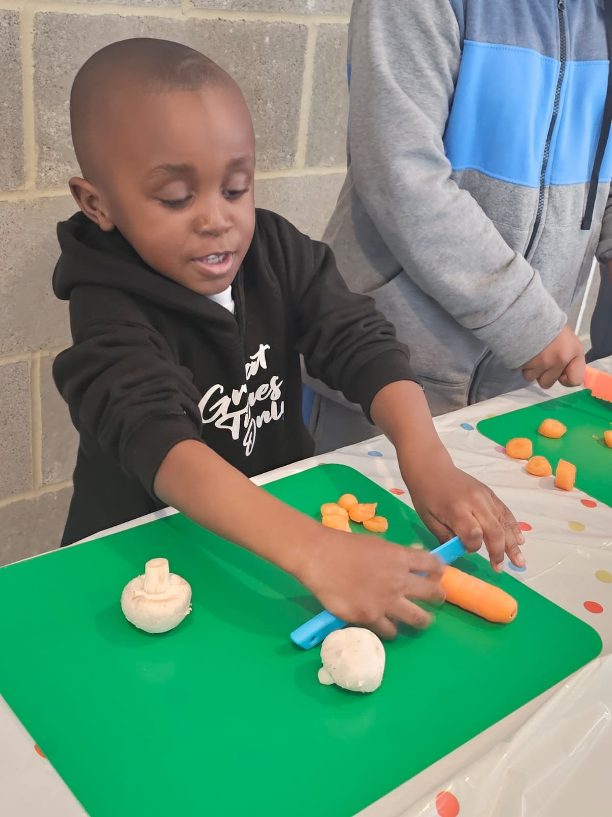 A young boy is cutting carrots on a green cutting board at a table with a festive tablecloth. There are also two white mushrooms on the board. Another person is standing nearby.