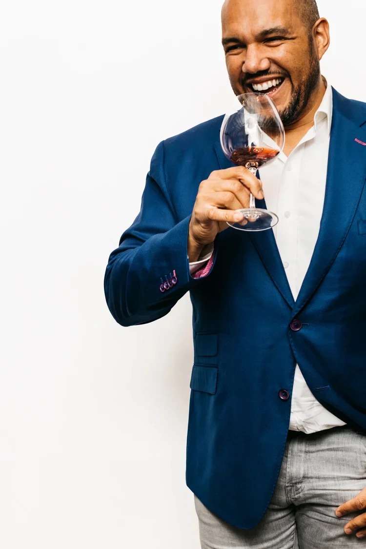 Boston wine expert TJ Douglas smiles and sips a glass of red wine while wearing a blue blazer.