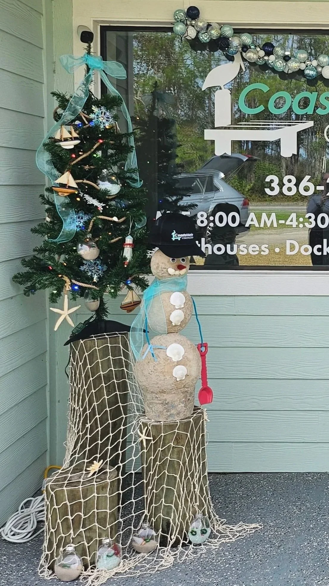 Decorative snowman made from stacked stones with a black hat, red nose, and buttons, standing next to a small decorated Christmas tree with blue ribbon and boat ornaments, placed on wood stumps covered with a fishing net, outside a building with glass window and signage.