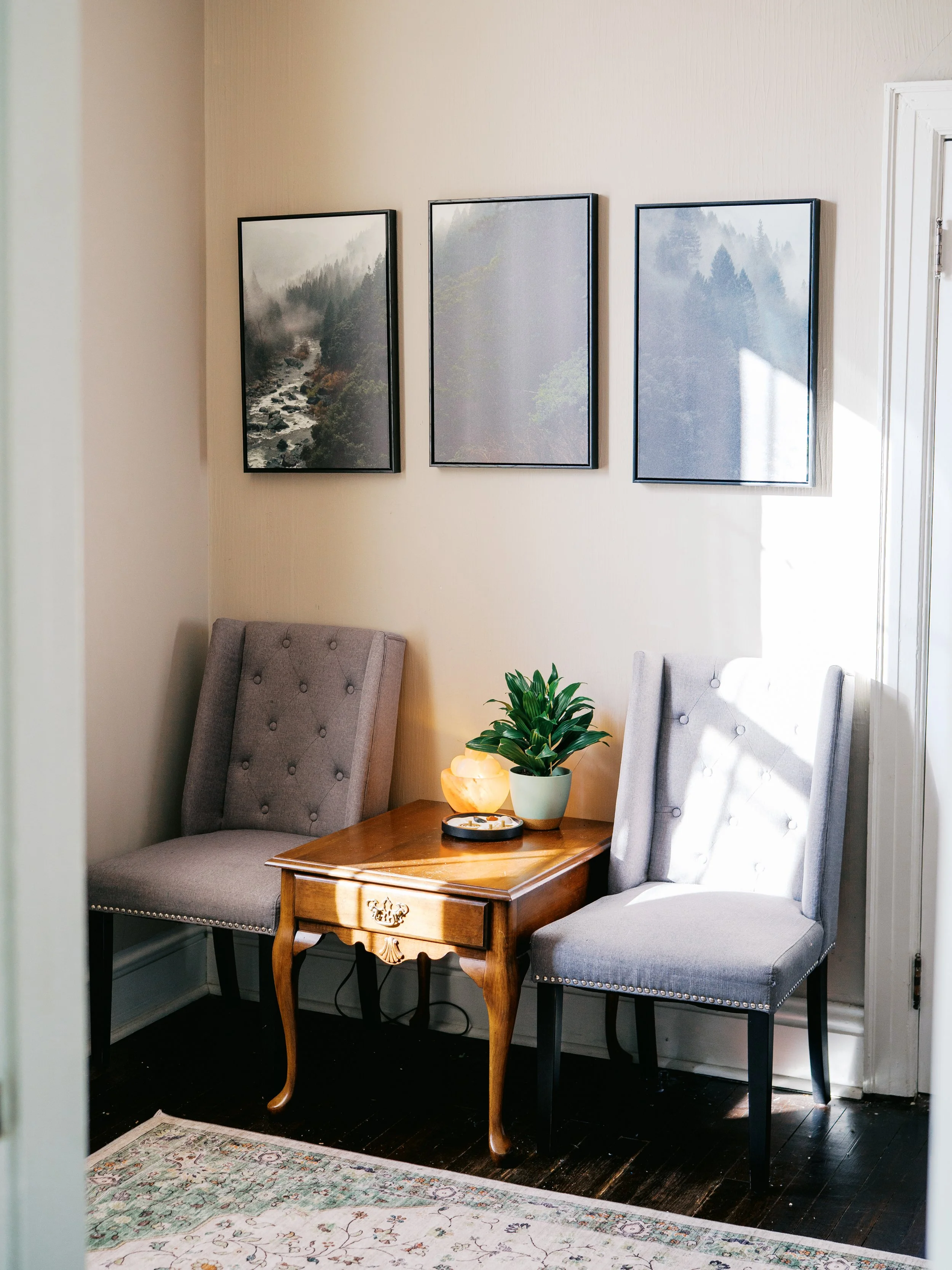 A cozy corner with two upholstered gray chairs, a wooden side table with a potted plant, a salt lamp, and an electric kettle. Wall art above features three landscape photos of foggy mountains and trees. Carpet and dark hardwood floor complete the setting.