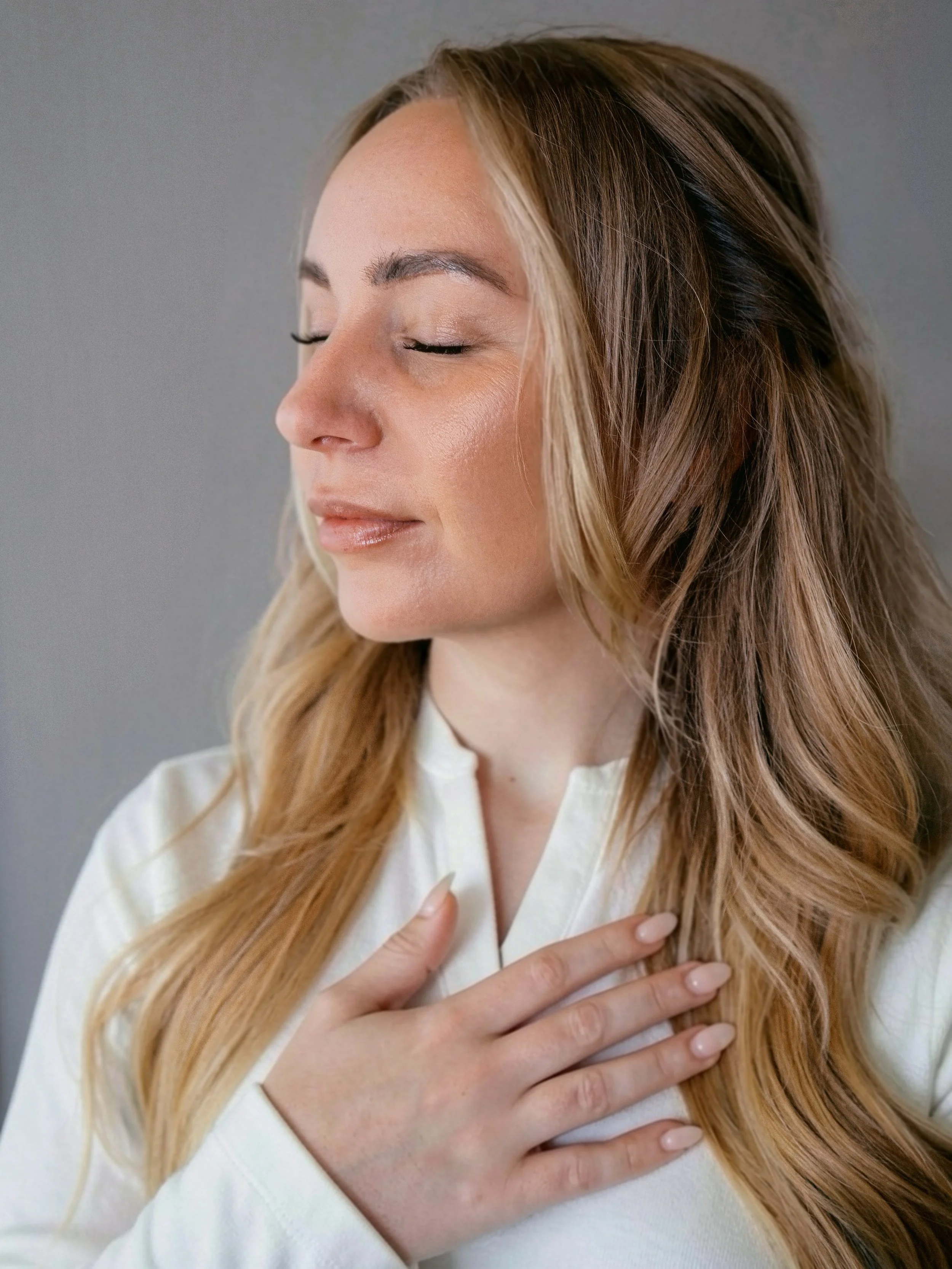 A woman with long, wavy blonde hair, closed eyes, and a peaceful expression, touching her chest with her right hand, wearing a white top.