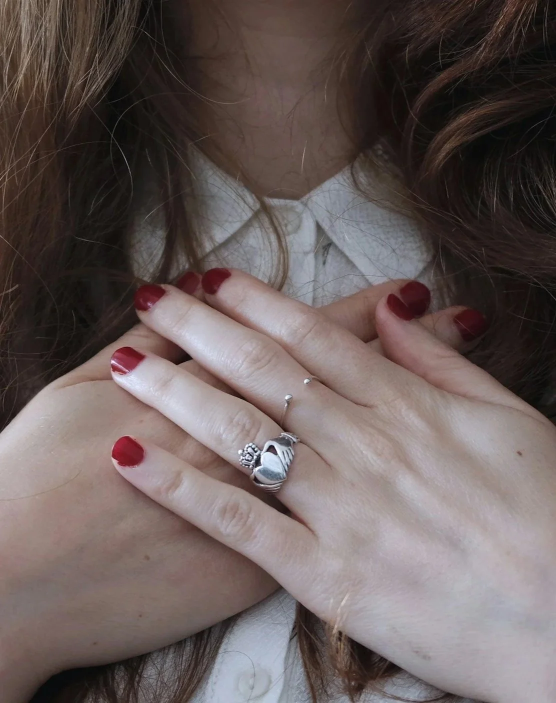 Person with red painted nails wearing multiple rings, including a heart-shaped ring, resting their hand on their shoulder. The person is wearing a white shirt and has long, wavy hair.