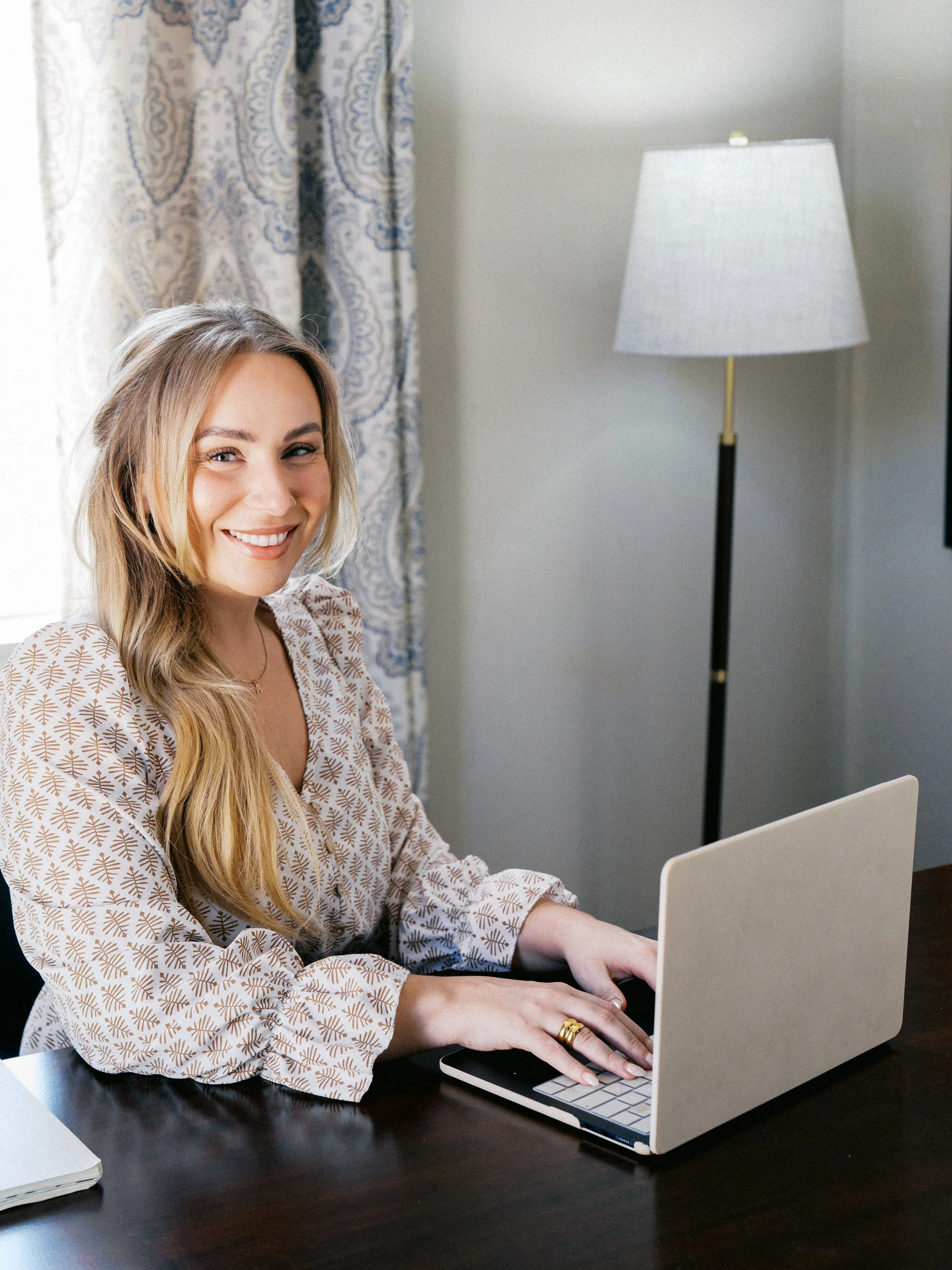 A woman with long blonde hair smiling while sitting at a dark wooden desk and typing on a silver laptop.