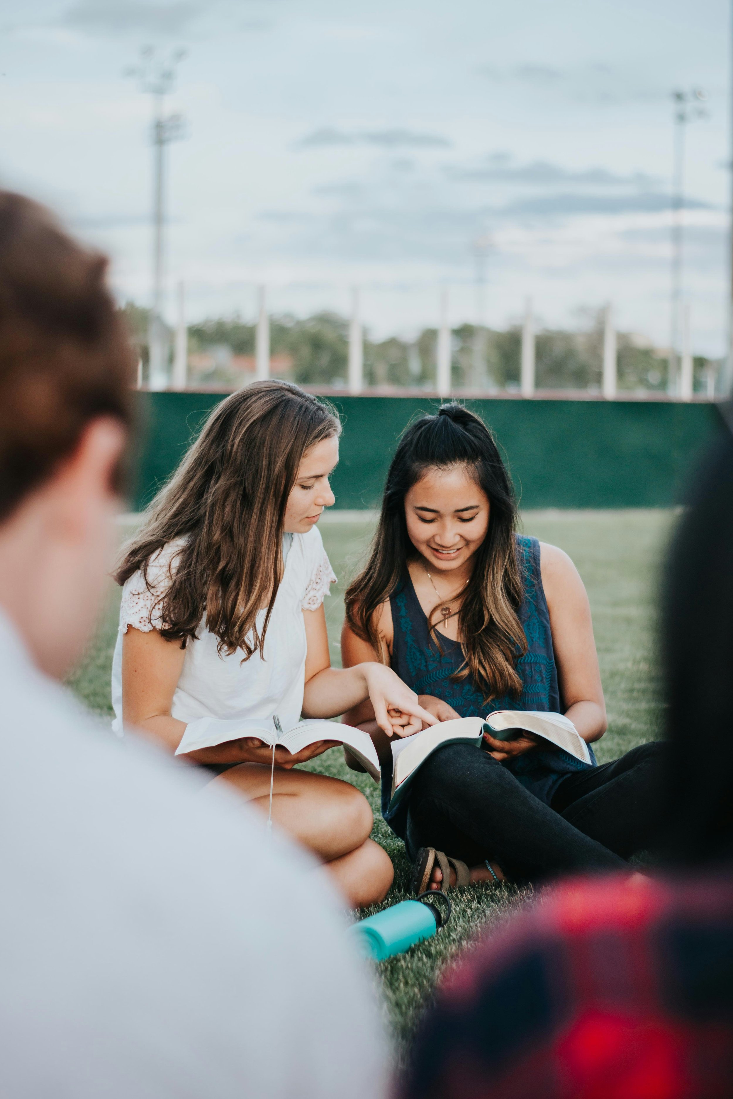 Two young women sitting on the grass, reading books and smiling, with other blurred individuals around them in a field with a green fence and cloudy sky in the background.