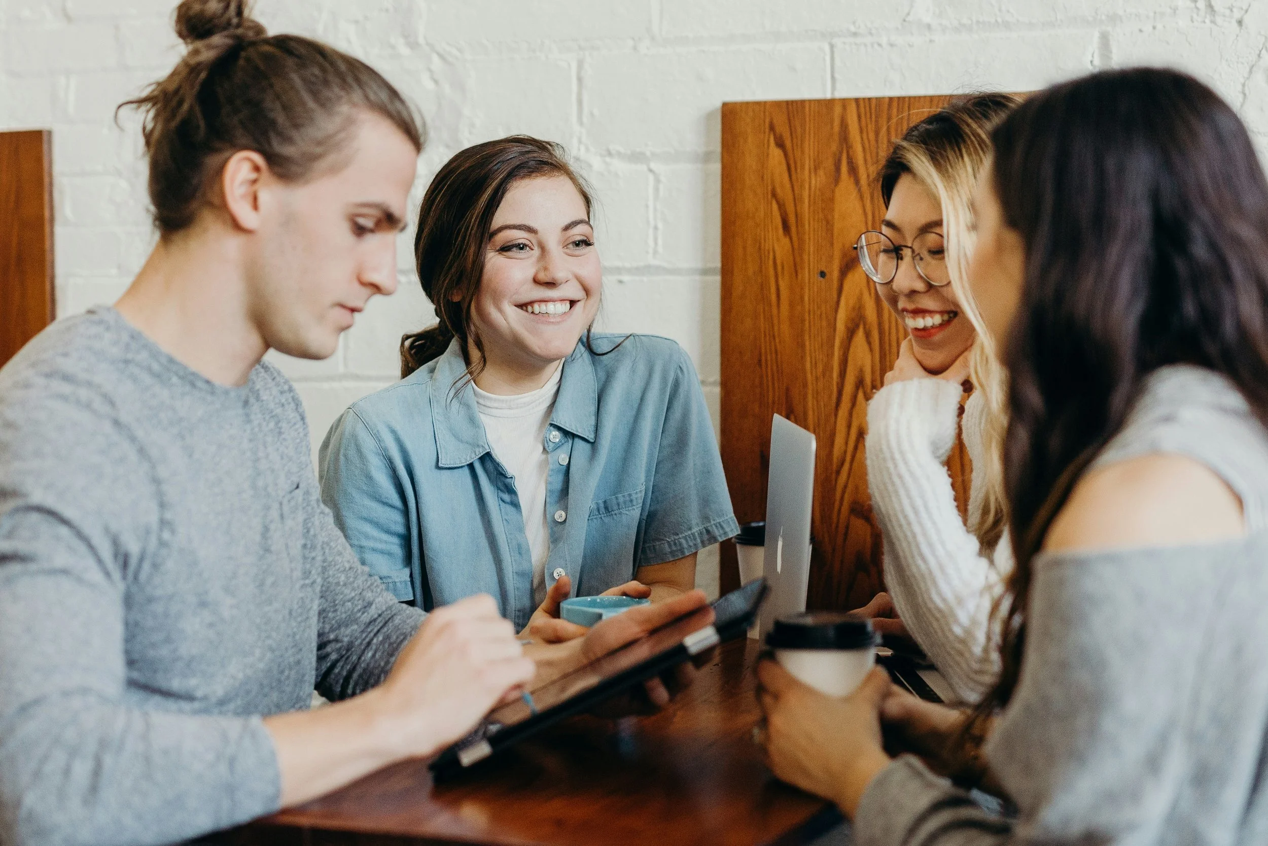 Four young adults sitting at a table in a cafe, engaging with a tablet, smiling and talking to each other.