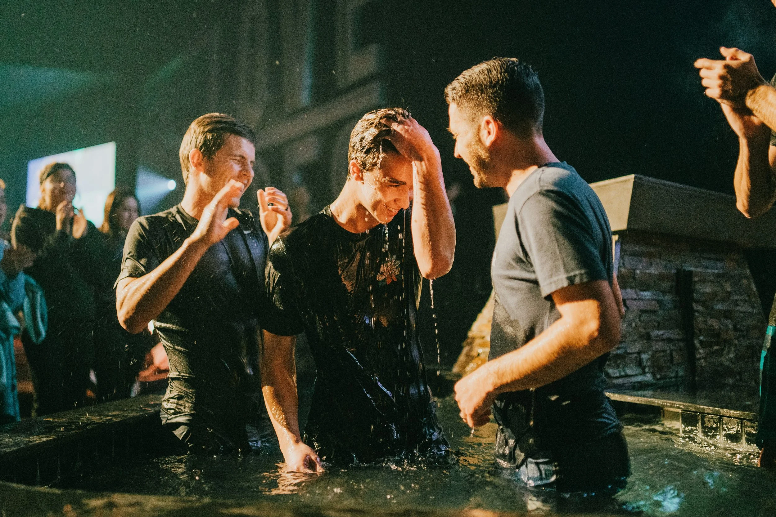 A group of people gathered around a baptismal pool, with a man being baptized, water pouring over his head, and others clapping and smiling in a church or religious setting.