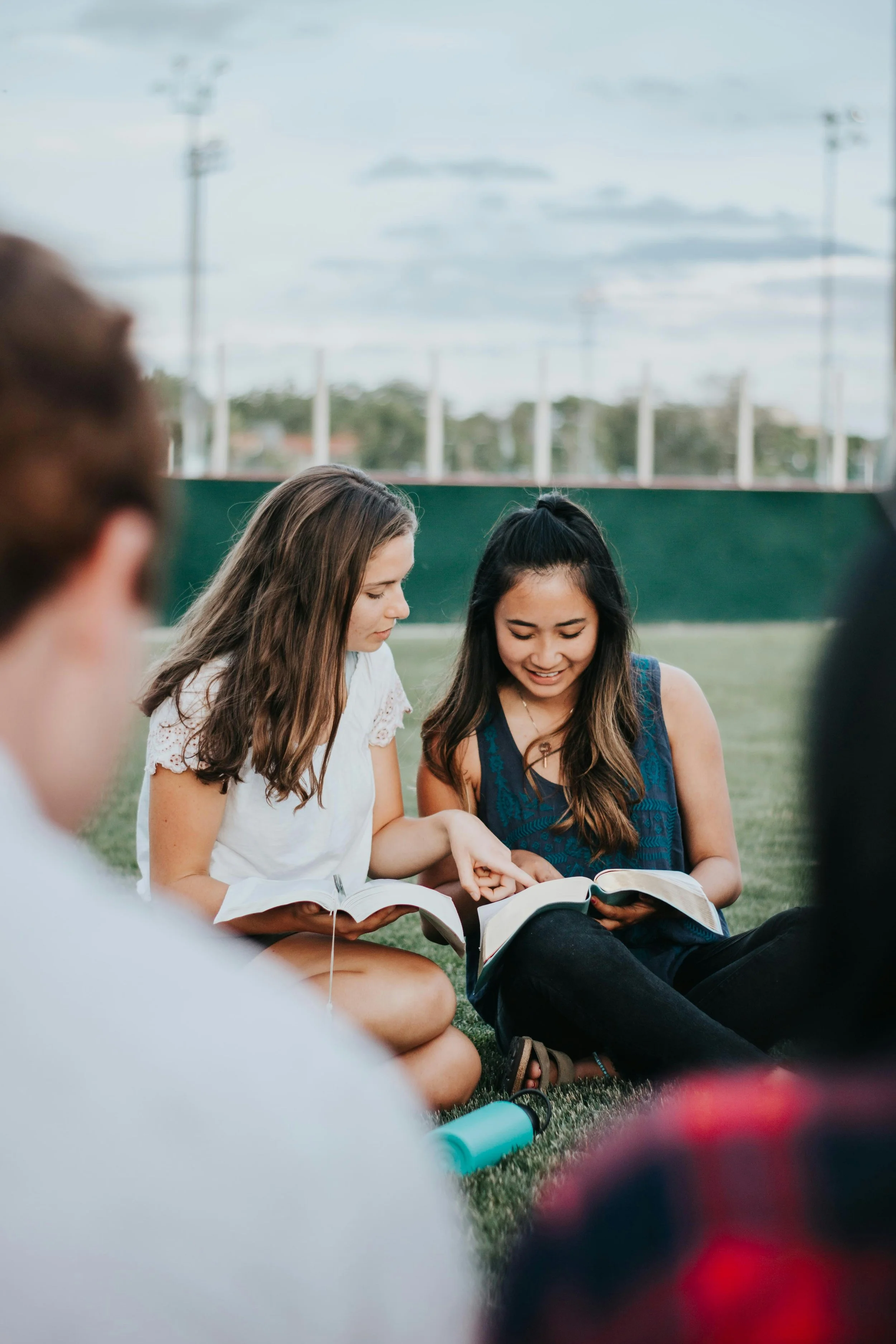 Two young women sitting on grass looking at and sharing a book together while others are partially visible around them, outdoors on a sports field.