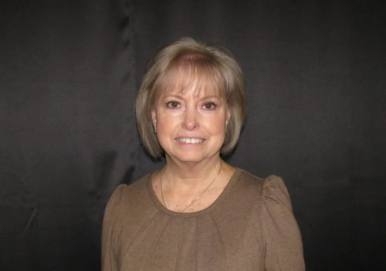 A smiling woman with short blonde hair, wearing a brown top, standing against a black background.