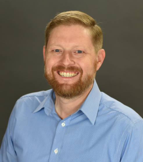 Portrait of a smiling man with red hair and beard wearing a light blue collared shirt against a gray background.