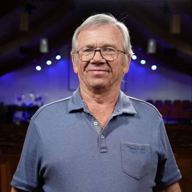 An elderly man with gray hair, glasses, wearing a blue polo shirt, standing in a dimly lit room with stage lights in the background.