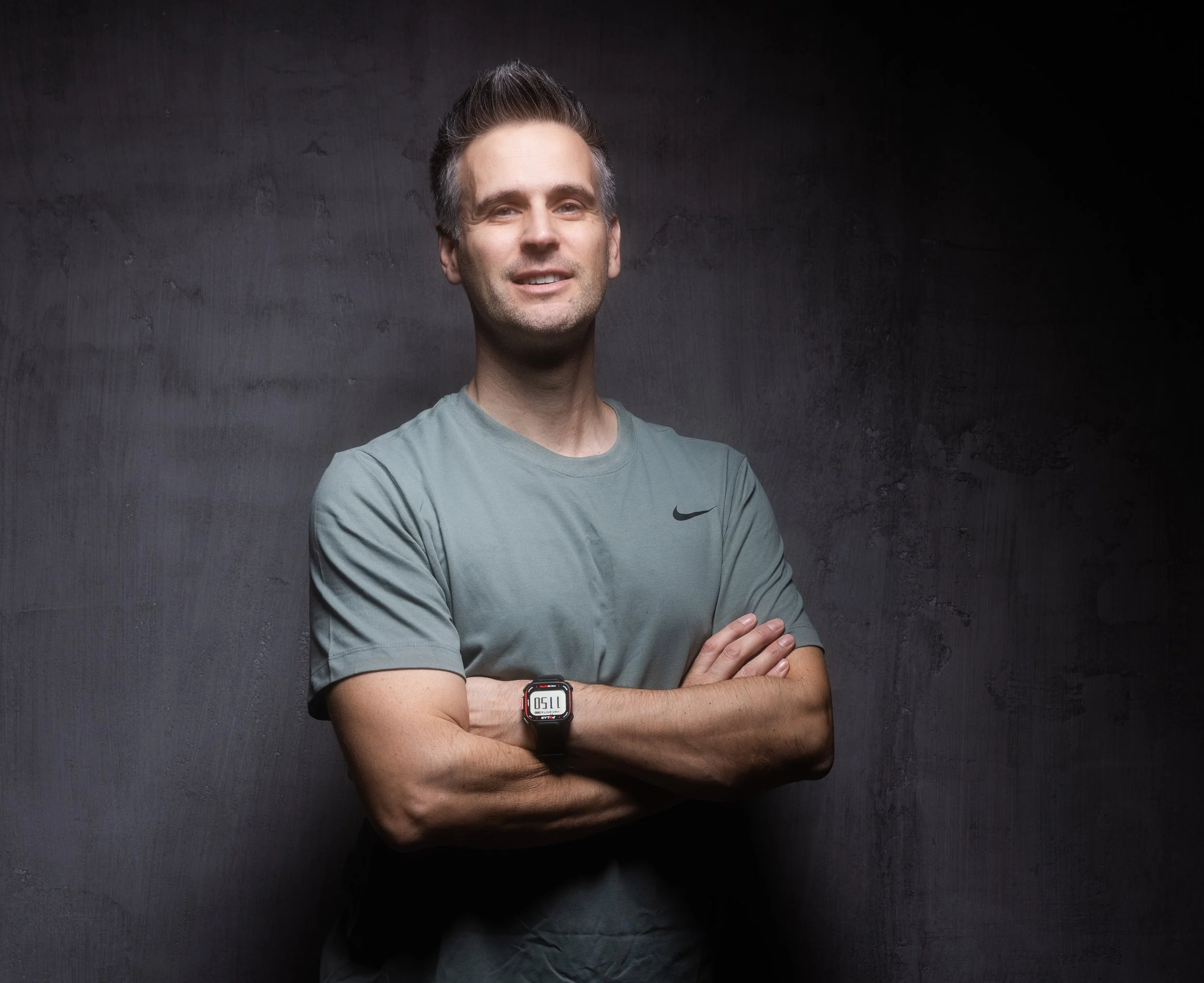 A middle-aged man with dark hair slicked back, wearing a blue short-sleeve sweater and beige pants, sitting on a bench against a beige wall with soft lighting and a shadow cast on the wall behind him.