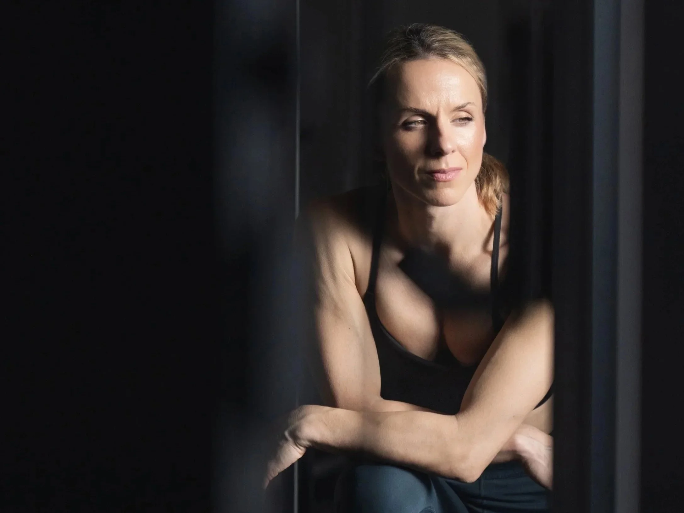 A woman with blonde hair wearing a blue sweatshirt, standing against a dark textured background, smiling with hands on hips.