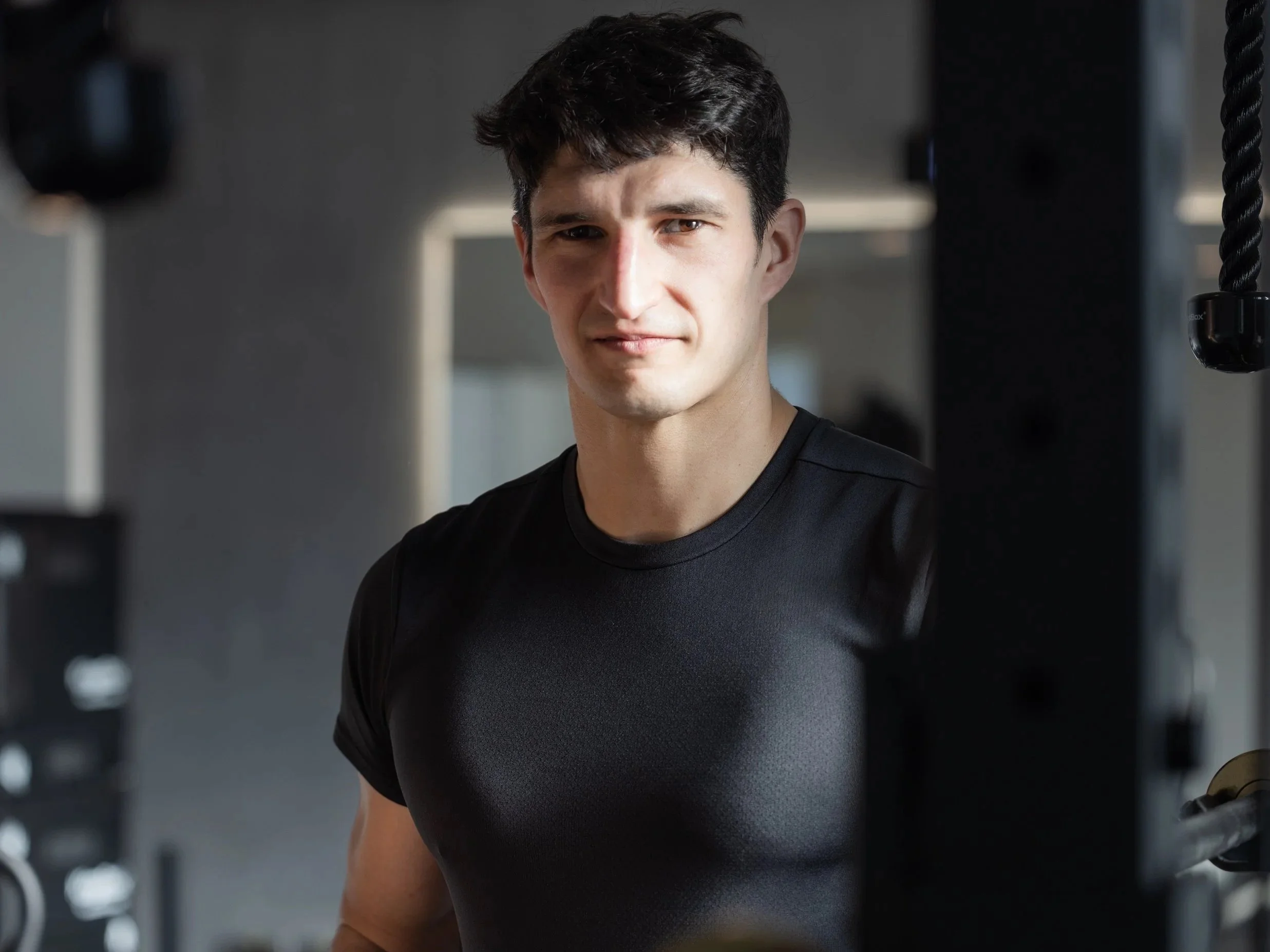 Woman with curly dark hair sitting on a bench, looking thoughtfully to the side, with sunlight casting shadows on the wall behind her in a modern room.
