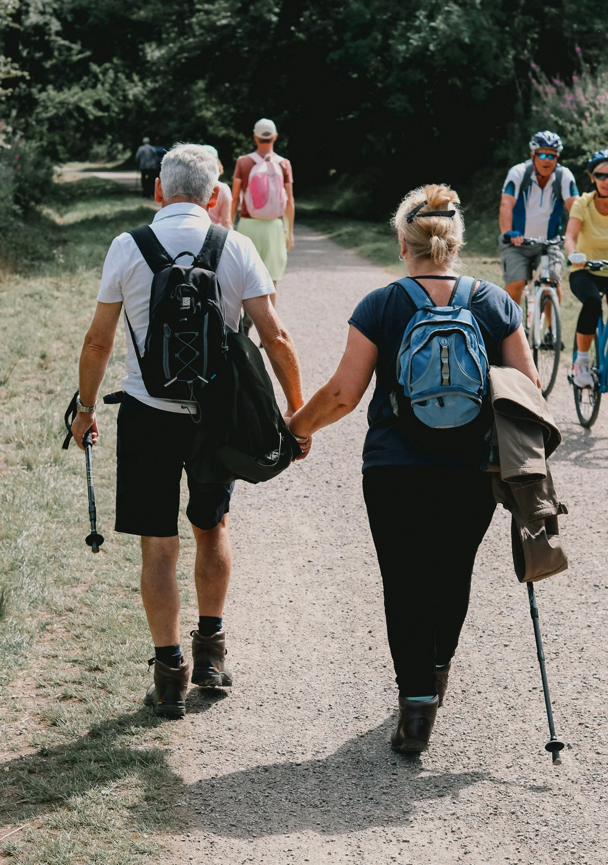 Older man and woman walking hand in hand on a gravel trail, carrying backpacks, outdoors, surrounded by trees, with cyclists and other pedestrians in the background.