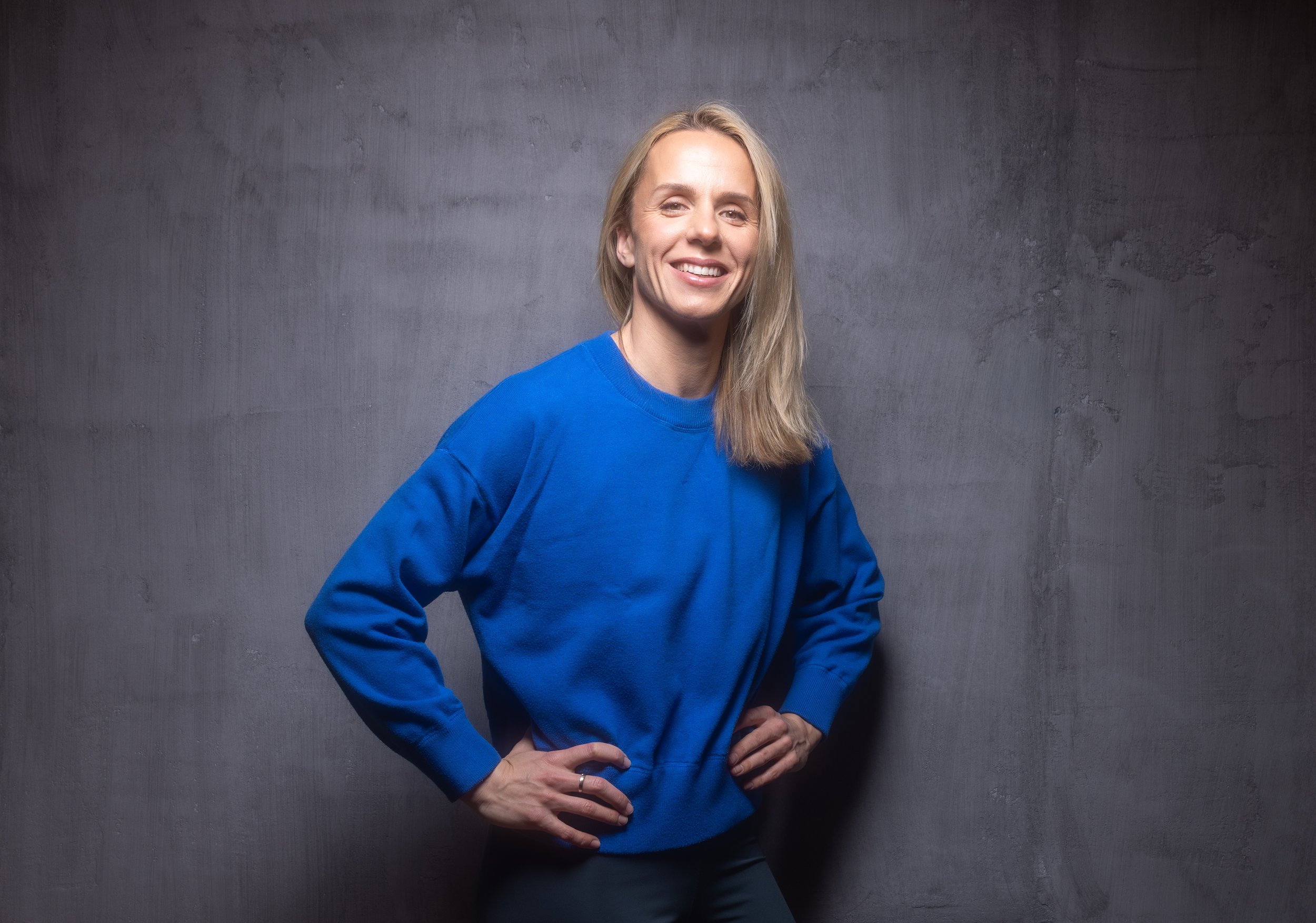 A woman with blonde hair wearing a blue sweatshirt, standing against a dark textured background, smiling with hands on hips.