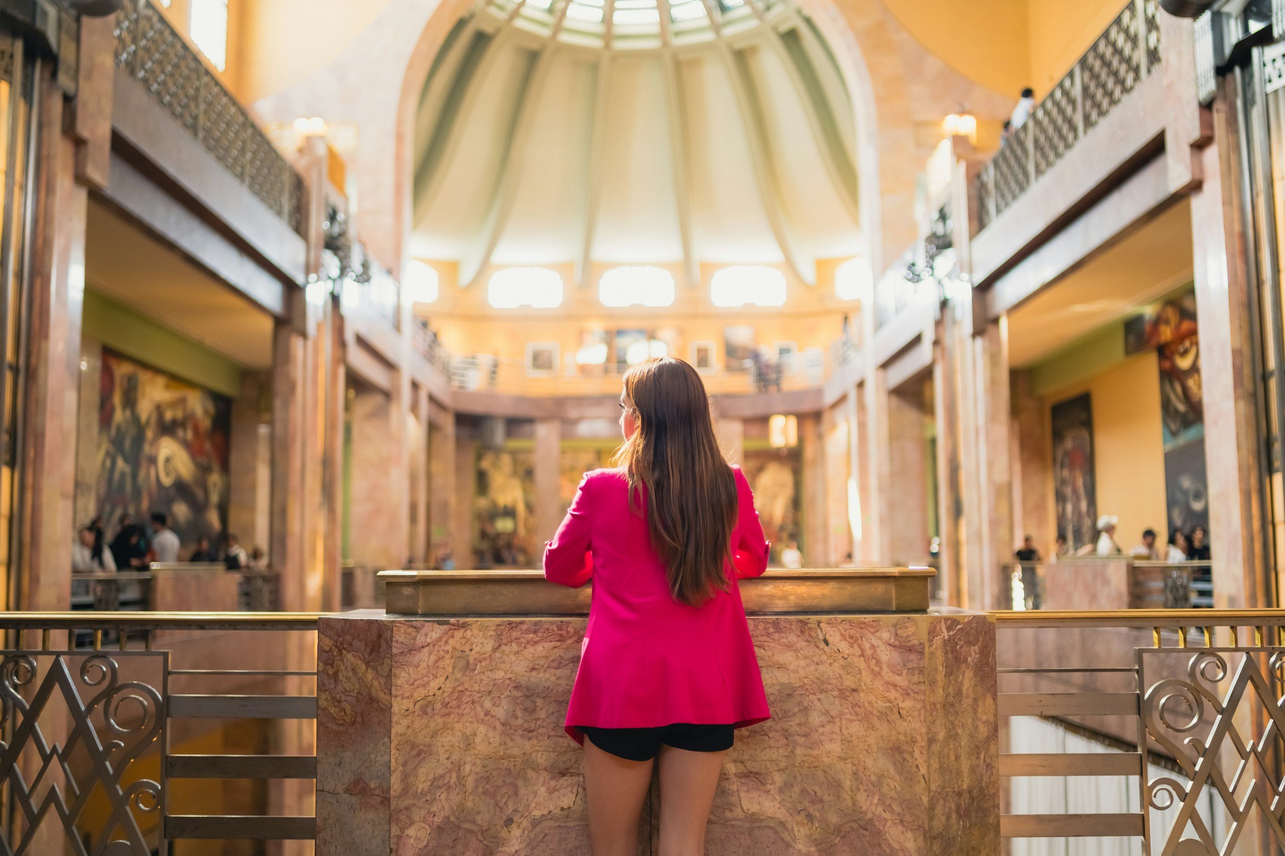 A woman in a pink blazer and black shorts standing at a marble counter inside a spacious, well-lit mall with high ceilings, balconies, and artwork.
