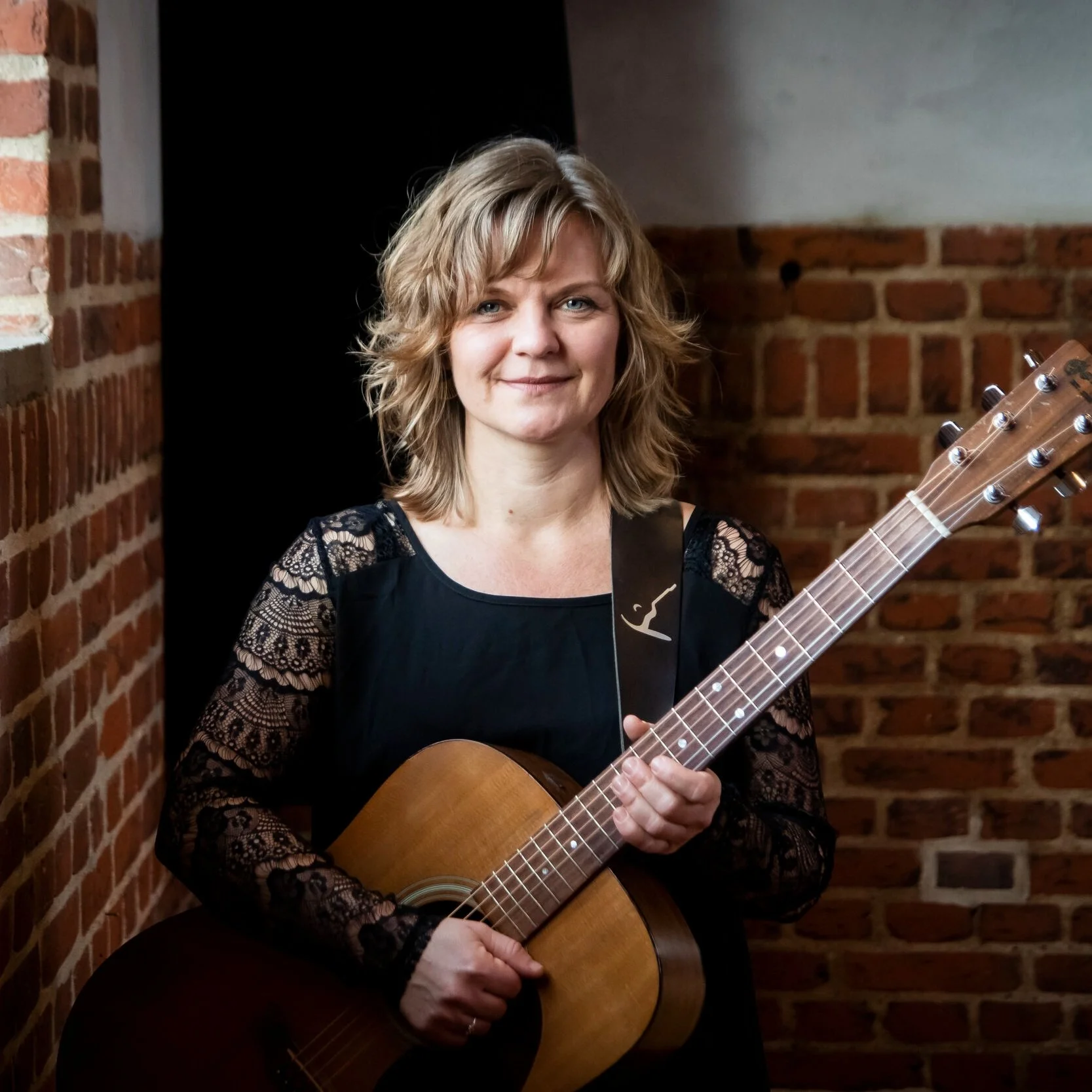 Kvindelig musiker med blond hår, sort bluse med blonde ærmer, spiller på en akustisk guitar i et rum med brune mursten.