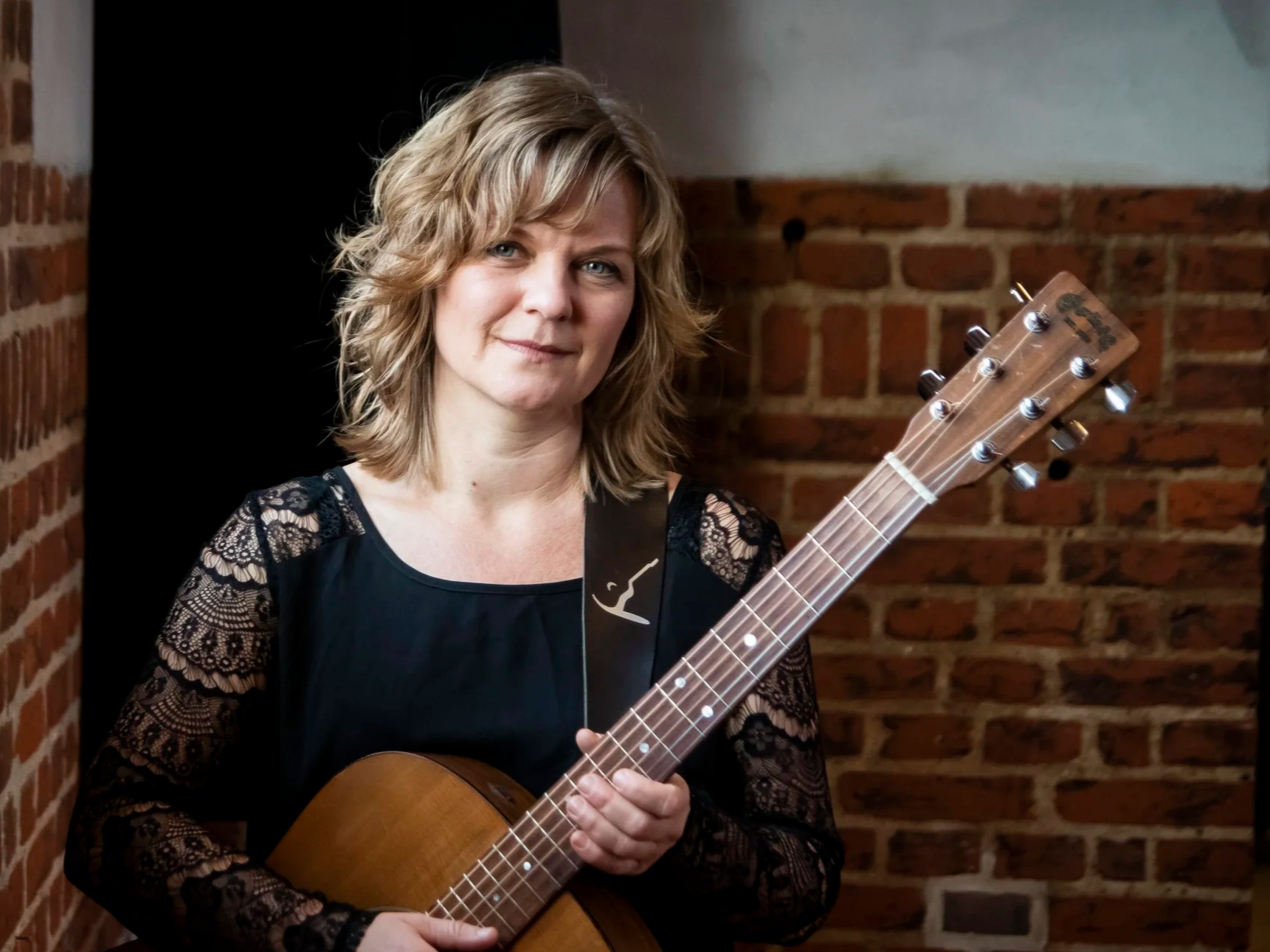 En kvindelig musikere holder en guitare foran en mur af røde mursten.