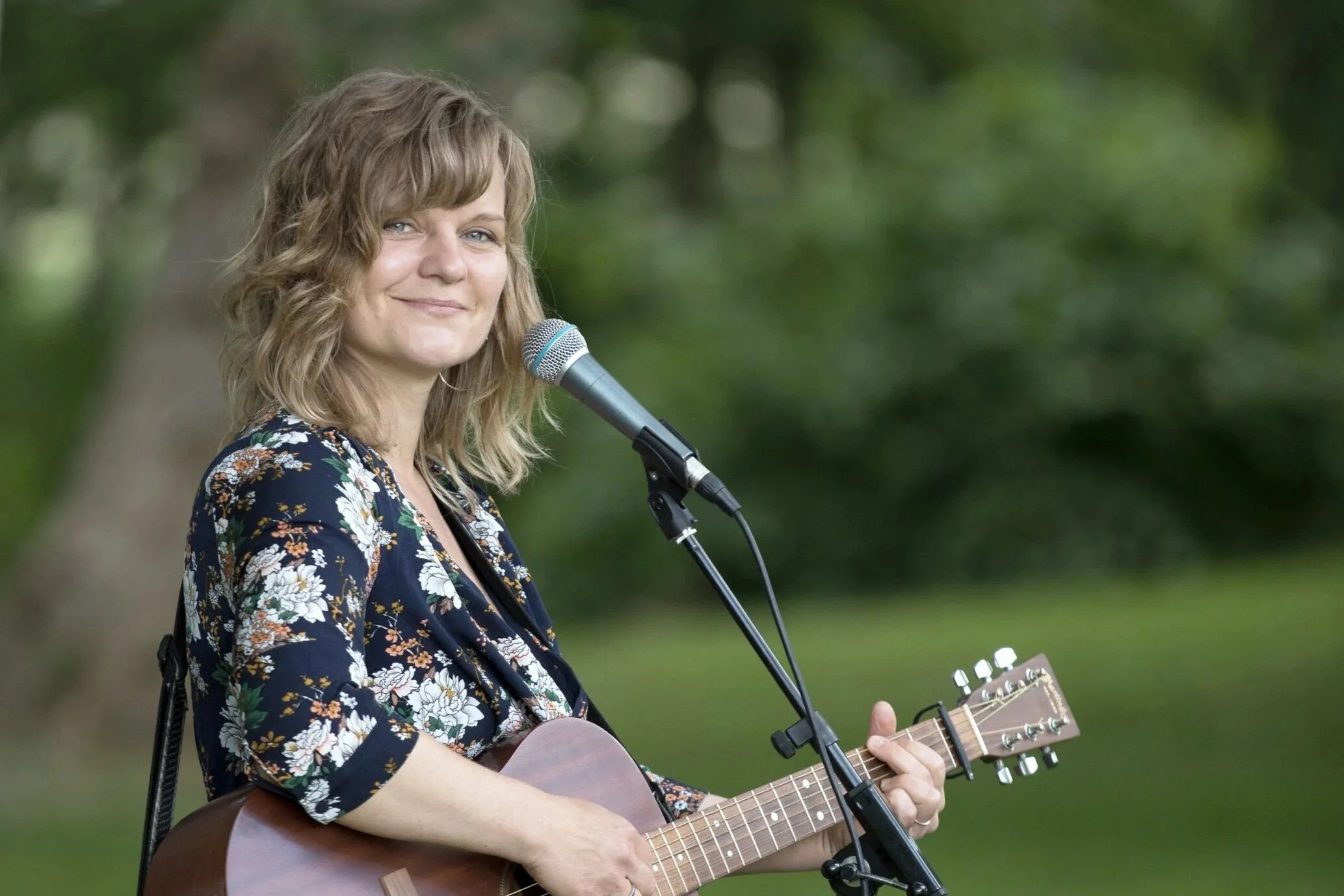 En kvinde med skulderlangt, bølget, blond hår spiller på akustisk guitar og synger foran en mikrofon udendørs med grønne træer og græs i baggrunden.