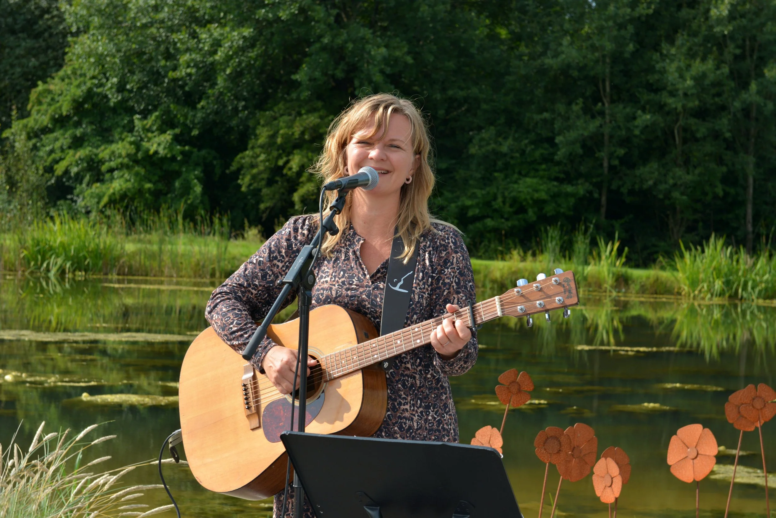 En kvinde spiller guitar og synger foran en sø med grønne træer i baggrunden.
