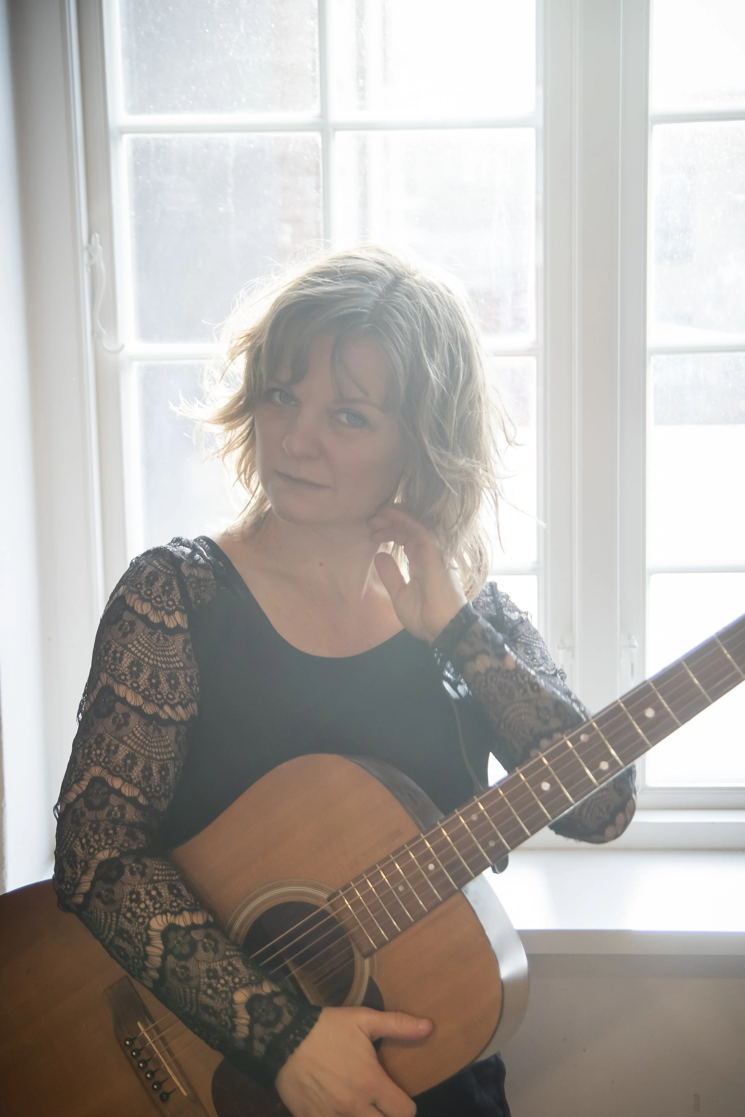 Kvindelig person med bølget lyst hår, der spiller guitar foran et vindue med flere små felter.