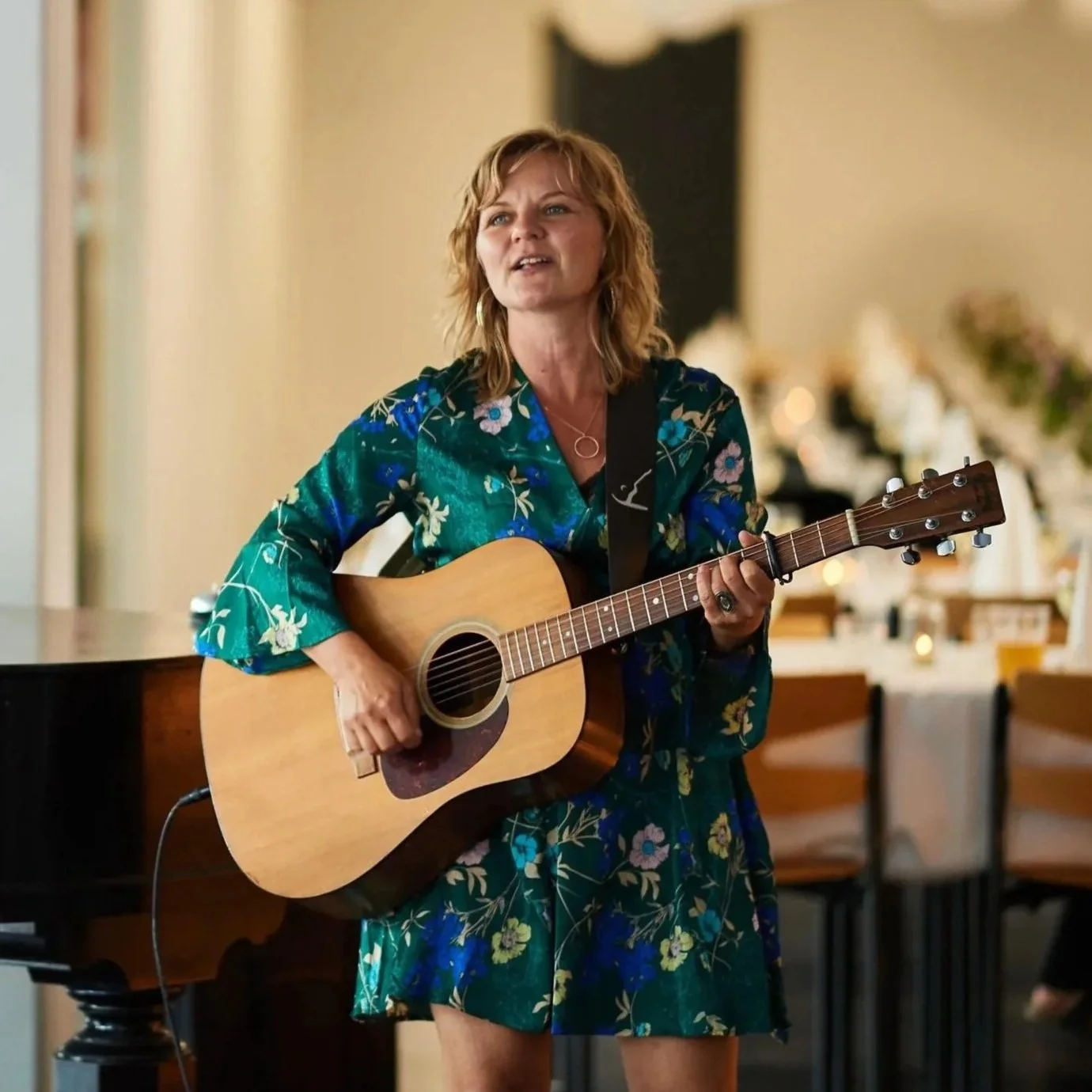 Kvinde spiller guitar i et spisested eller festlokale, iført en grøn blomstret kjole.