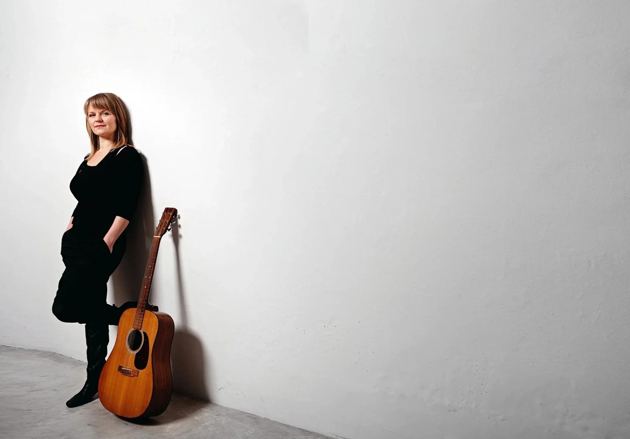 En kvinde med blond hår iført sort tøj, står imod en hvid væg med en guitar ved sin side.