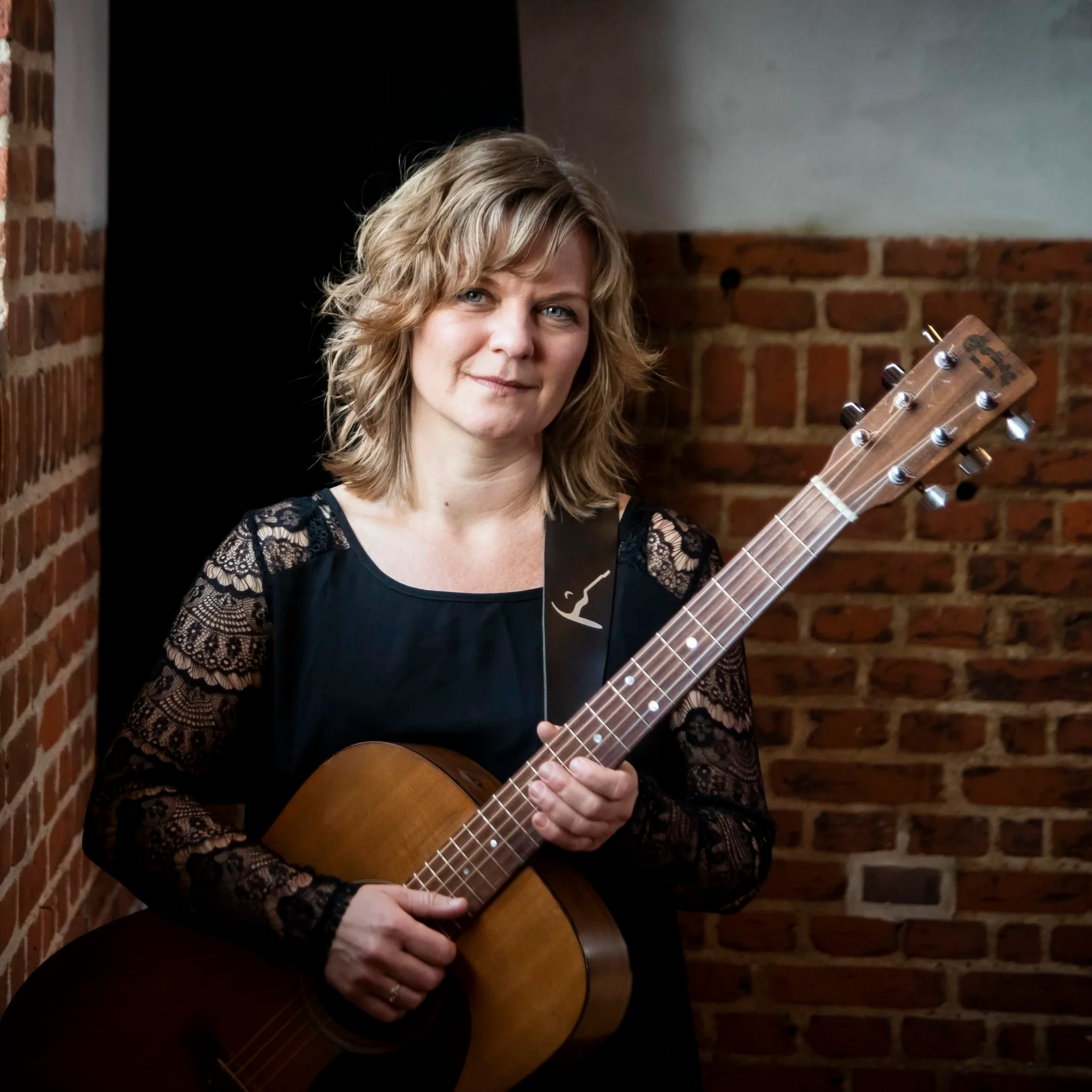 Kvinde med krøllet blond hår, klædt i sort med blonde puffer på ærmerne, holder en akustisk guitar foran en murstensvæg.
