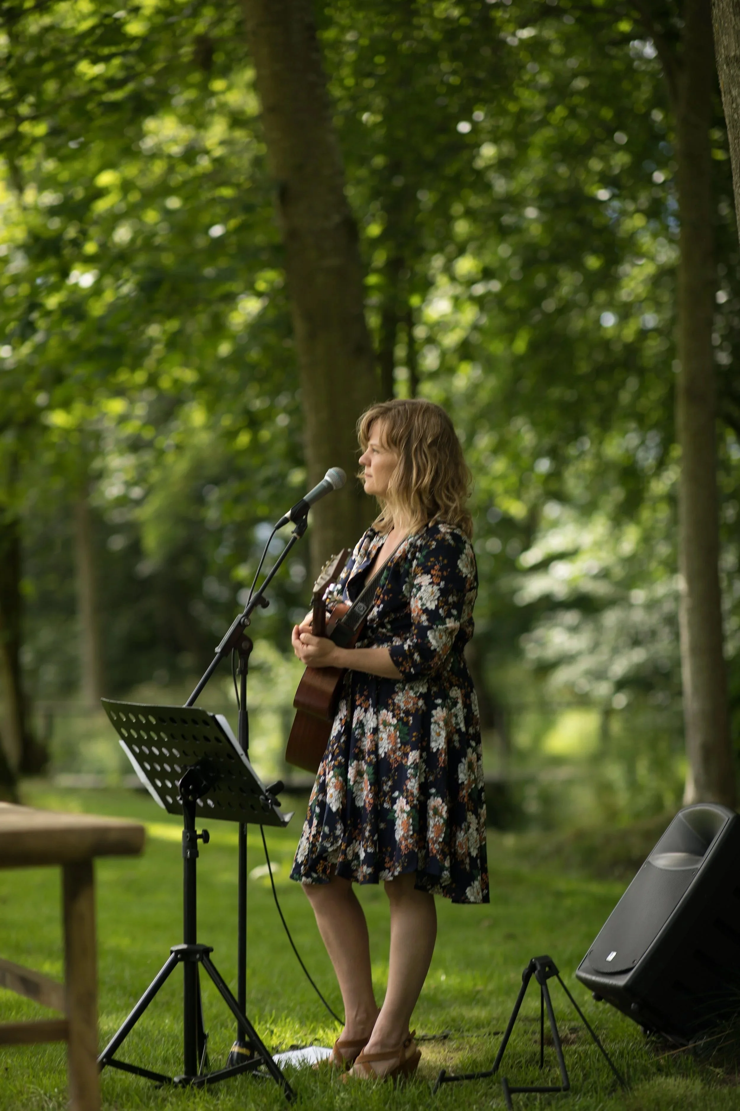 En kvinde står på græs i en skov, spiller guitar og synger foran en mikrofon, med en musikanlæg ved siden af.