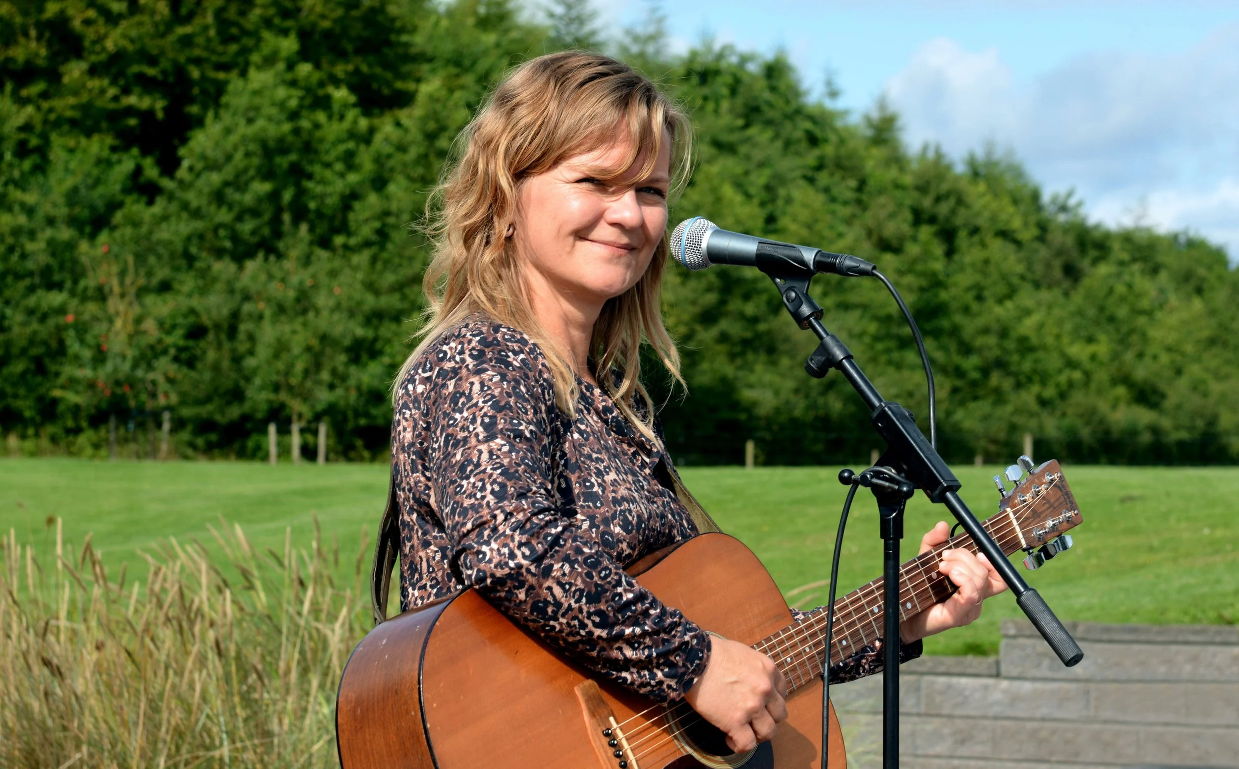 Kvinde med blond hår spiller på guitar udendørs, ved microphone, grøn natur og blå himmel i baggrunden.