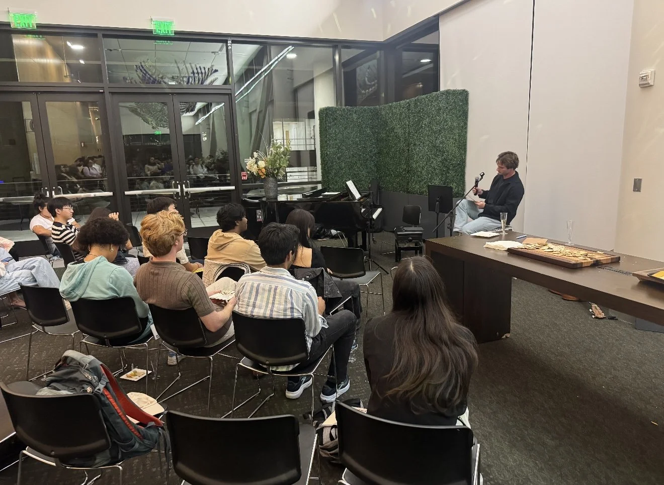 A man sitting on a chair and holding a microphone in front of an audience in a large indoor venue. The audience is seated facing him, some holding papers or food. There is a black piano with a floral arrangement nearby. The setting looks like a talk or performance event with people listening attentively.